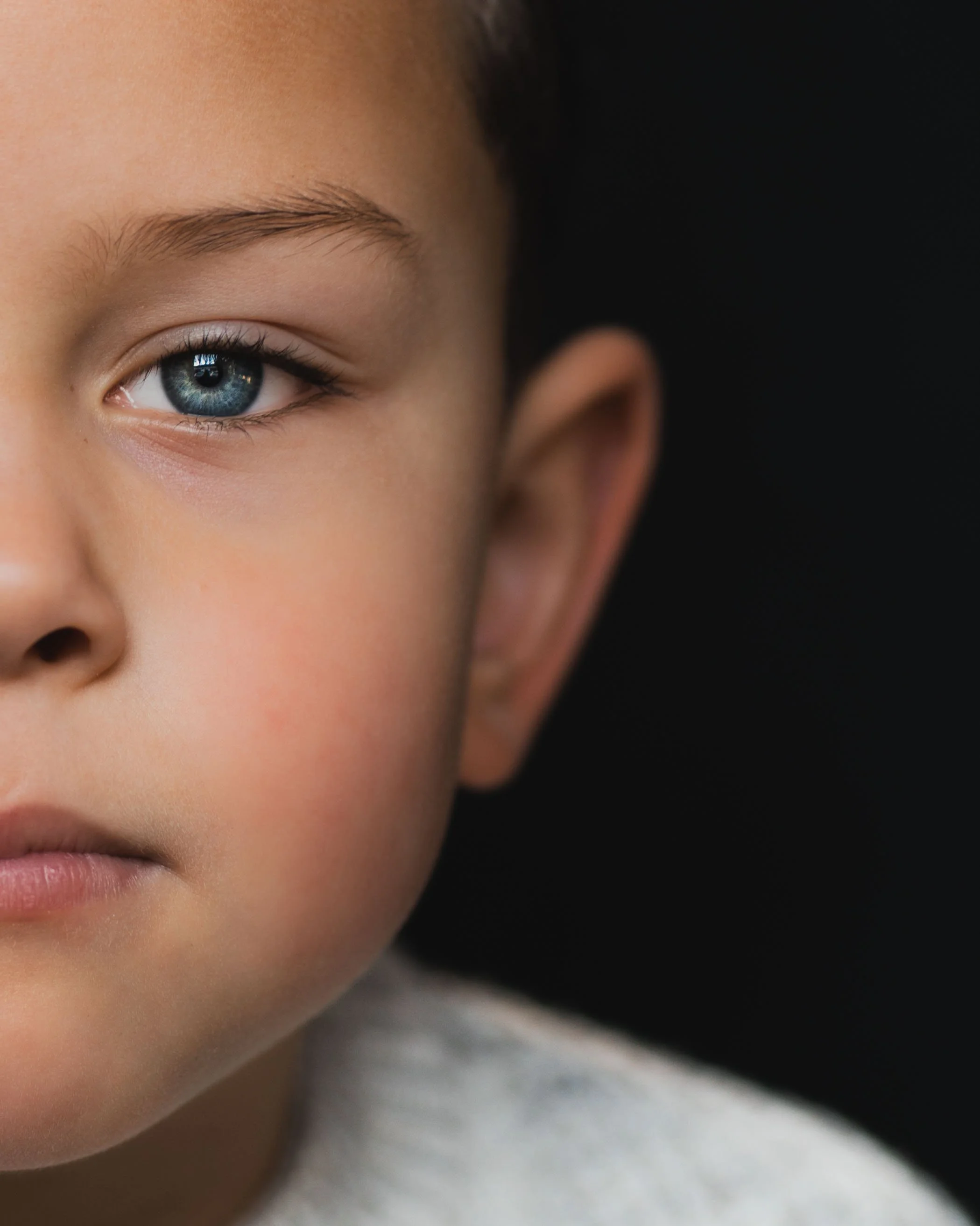 Close-up of a child's face showing one blue eye, part of the nose, lips, and ear against a dark background.