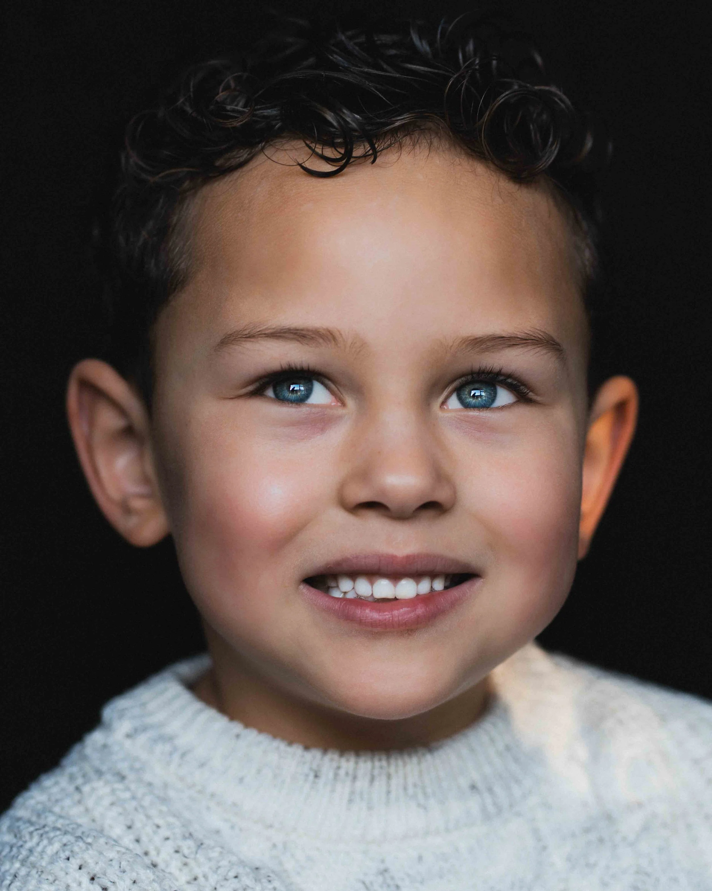 Close-up portrait of a young boy with curly dark hair, blue eyes, and a big smile, wearing a white knitted sweater against a black background.