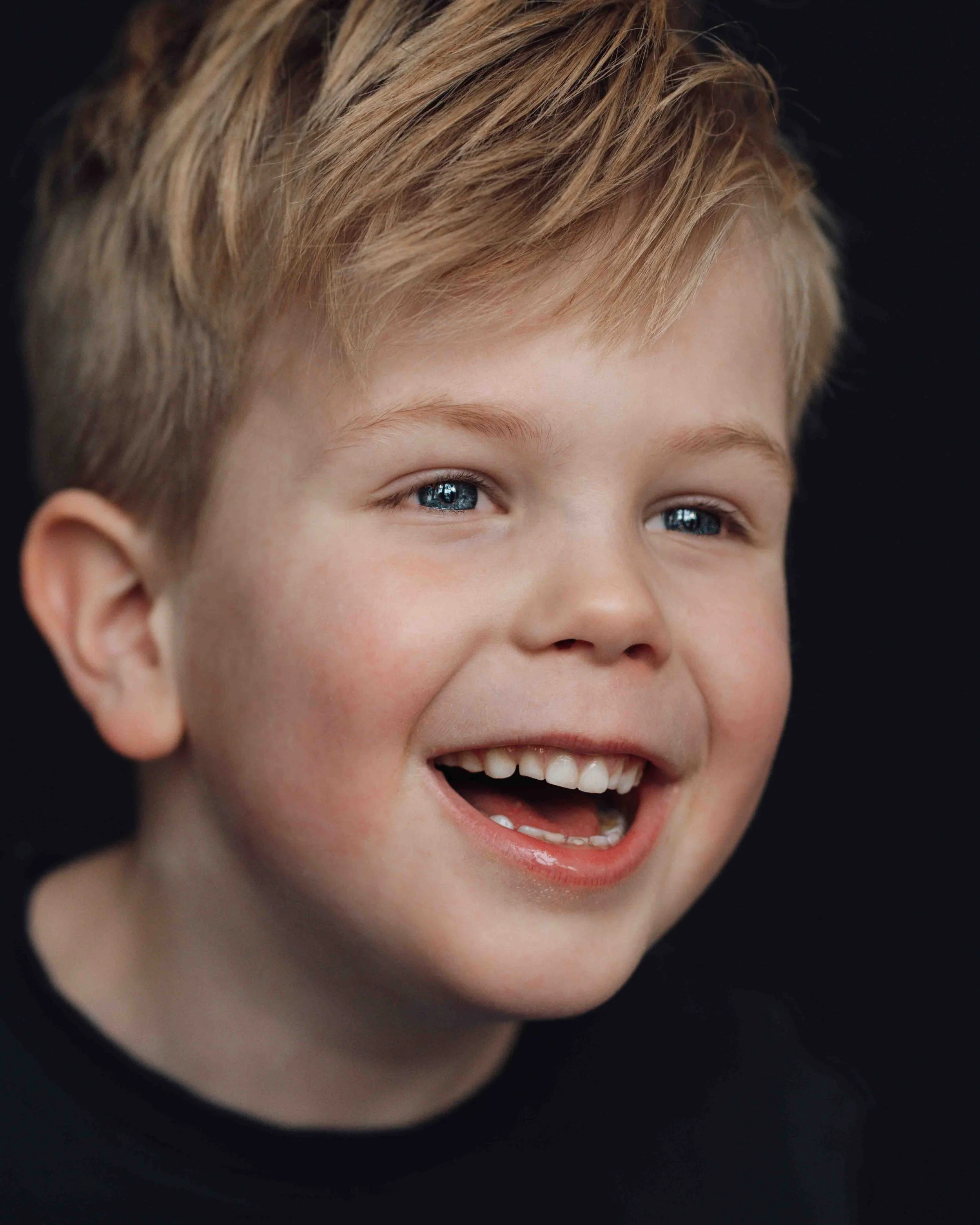 A young boy with blonde hair and blue eyes smiling widely, showing his teeth, against a dark background.