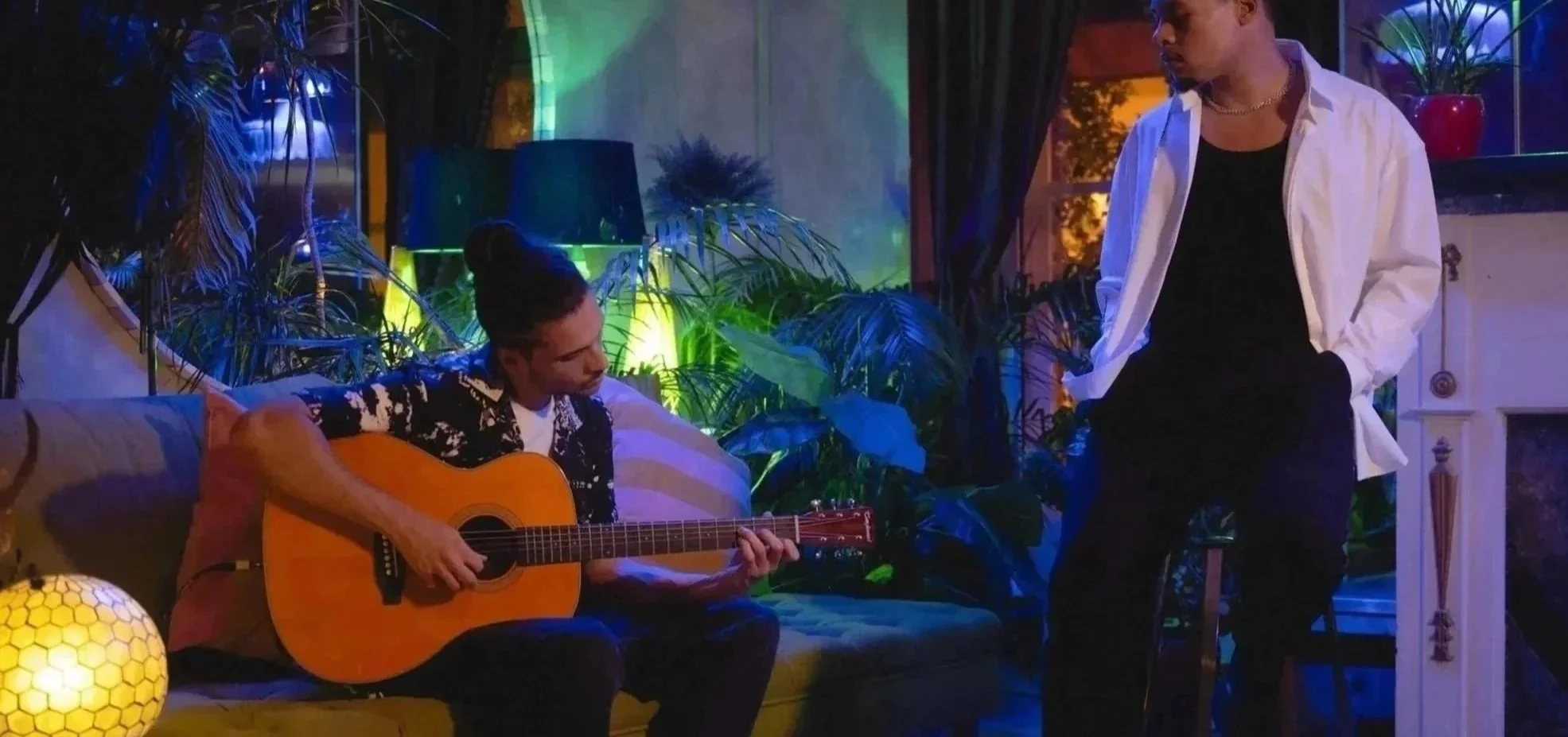 Two young men in a cozy indoor space with lush green plants. One is sitting on a futon playing an acoustic guitar, wearing a black and white patterned shirt. The other is standing, wearing a black shirt and white oversized shirt, looking at the guitarist. The room has colorful ambient lighting, lamps, and a white fireplace with a red plant pot on top.