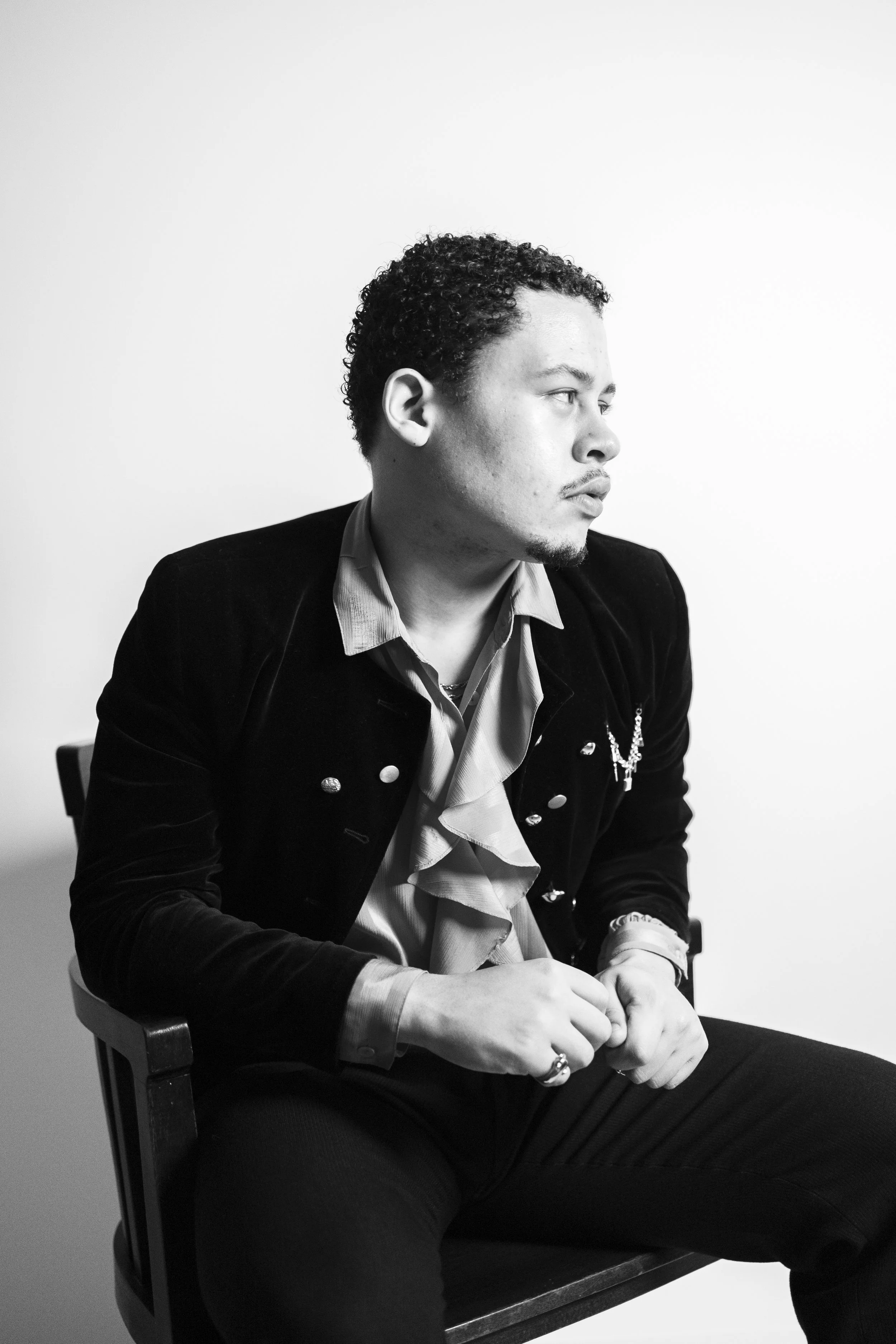 Black and white portrait of a man with curly hair, sitting on a chair, looking to the right, wearing a blazer over a ruffled shirt with a necklace, against a plain background.