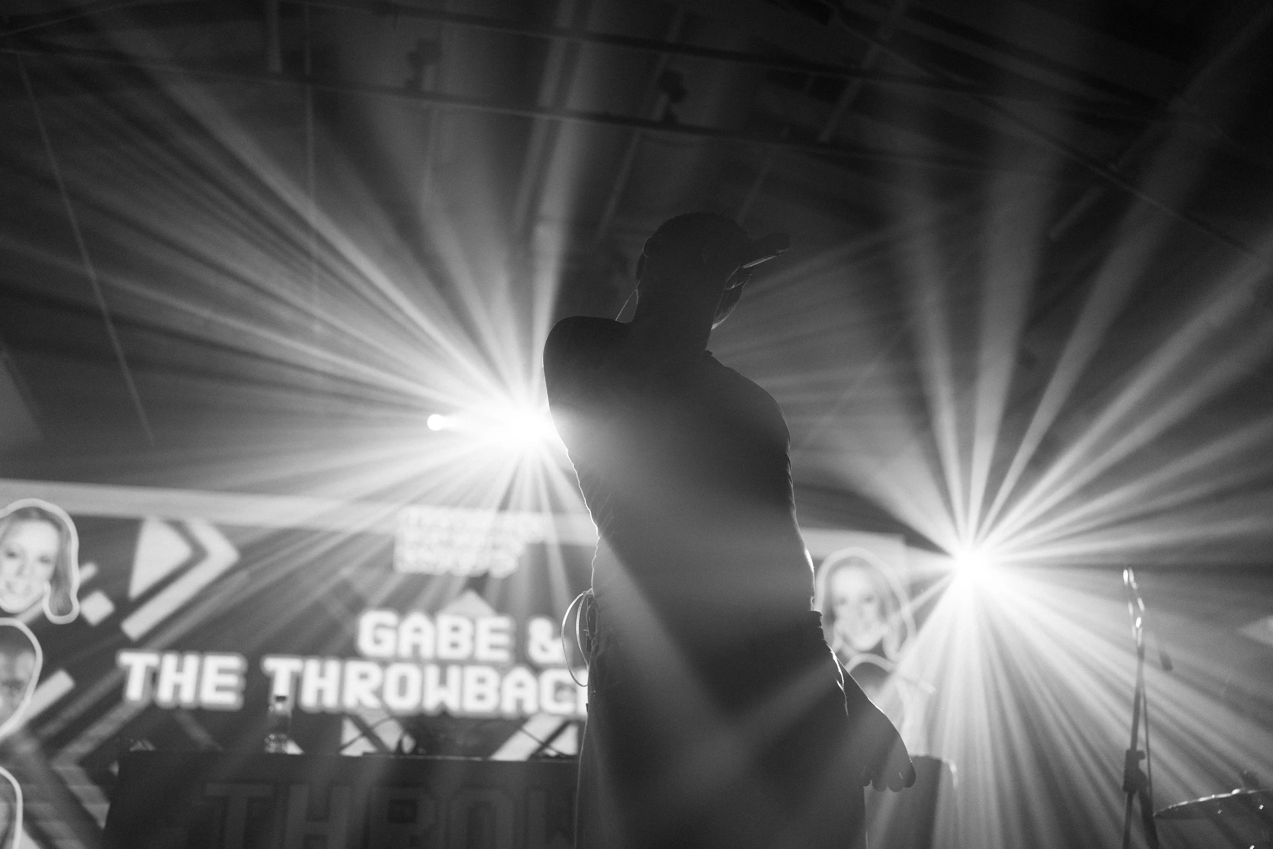 A person in a hat and glasses on stage with bright lights shining behind them, creating a silhouette effect, while a screen displays the text 'GABE & THE THROWBACK' in the background.