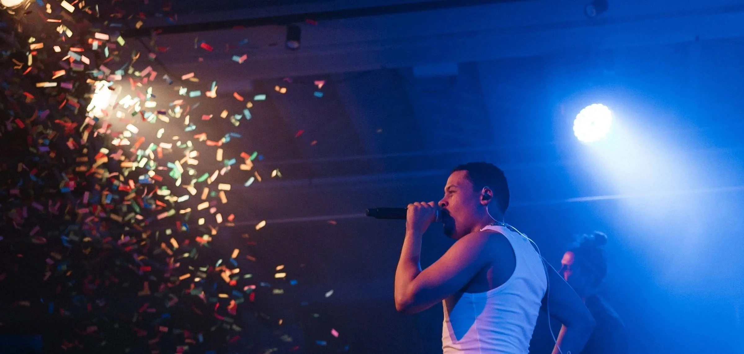 Performer singing into microphone on stage with colorful confetti and stage lights.