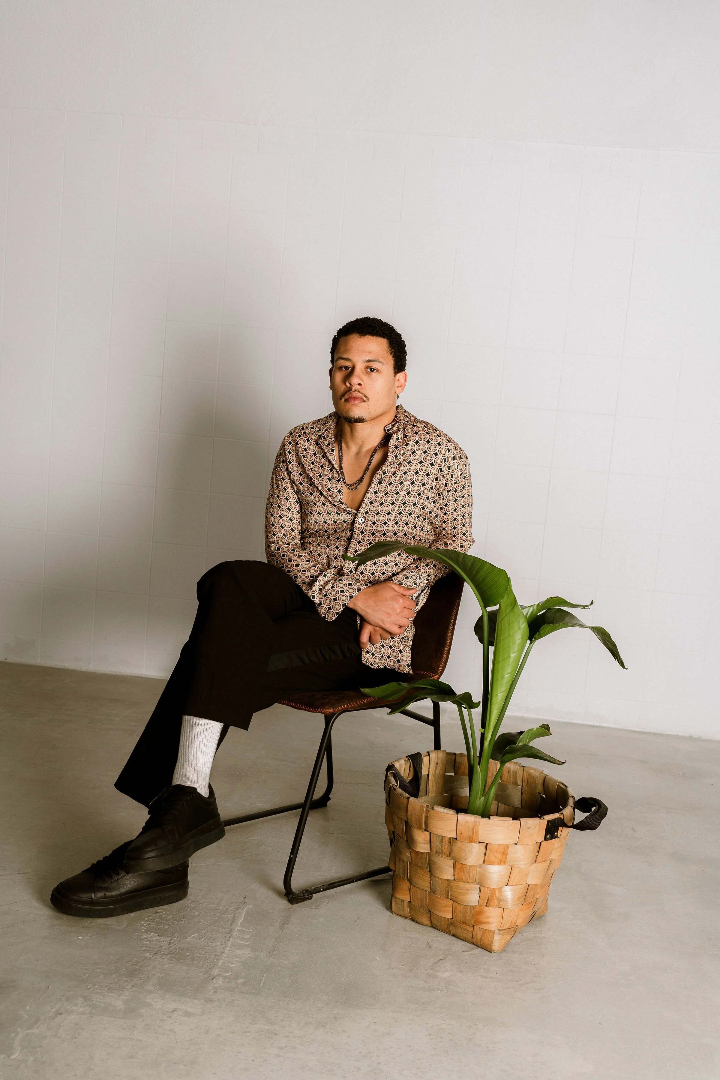 A young man with curly hair sitting on a chair with crossed legs, wearing a patterned shirt, black pants, black shoes, white socks, and a necklace, next to a potted plant in a woven basket against a plain white wall.