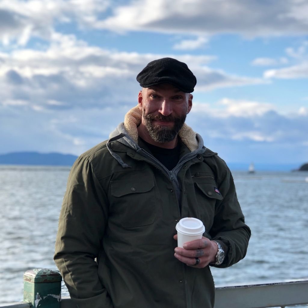 A man with a beard in a black cap stands on a boat or dock near water, holding a to-go coffee cup, with mountains, a boat, and a cloudy sky in the background.