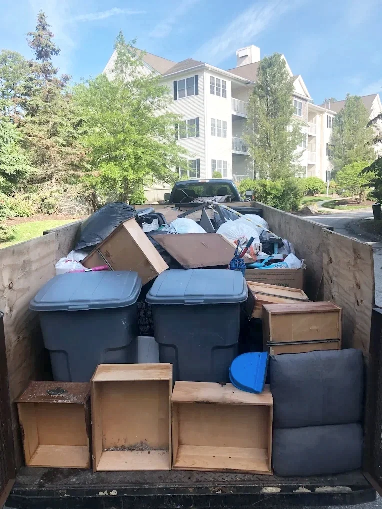 A trailer filled with various household items, including storage bins, wooden furniture, boxes, and miscellaneous debris, parked in front of a residential apartment complex.