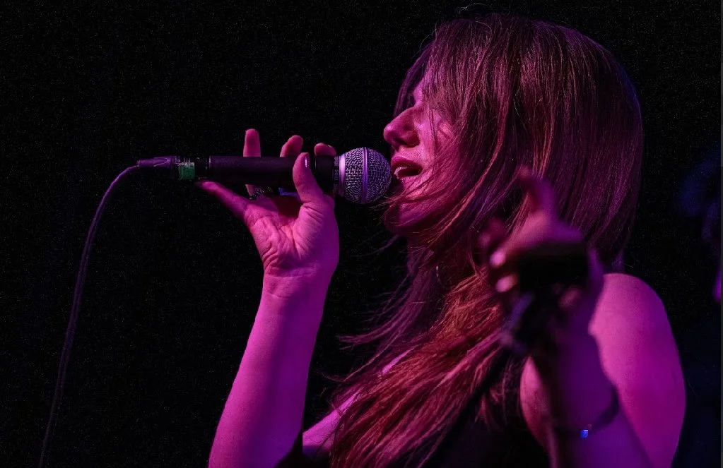 A woman singing into a microphone on stage with purple and pink lighting.