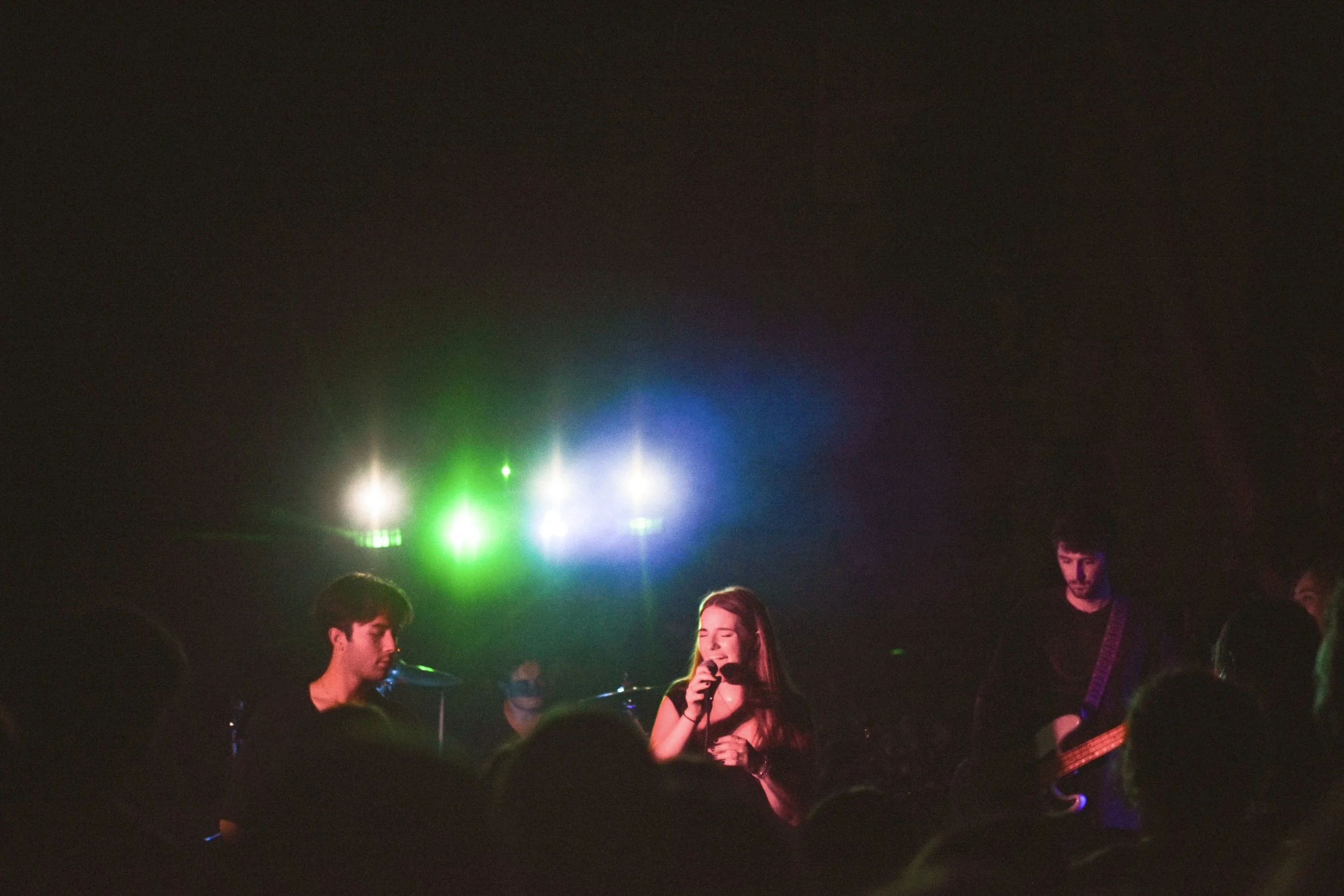 A young woman singing into a microphone on stage, flanked by two musicians playing guitar and bass, with colorful stage lights overhead, during a live concert.