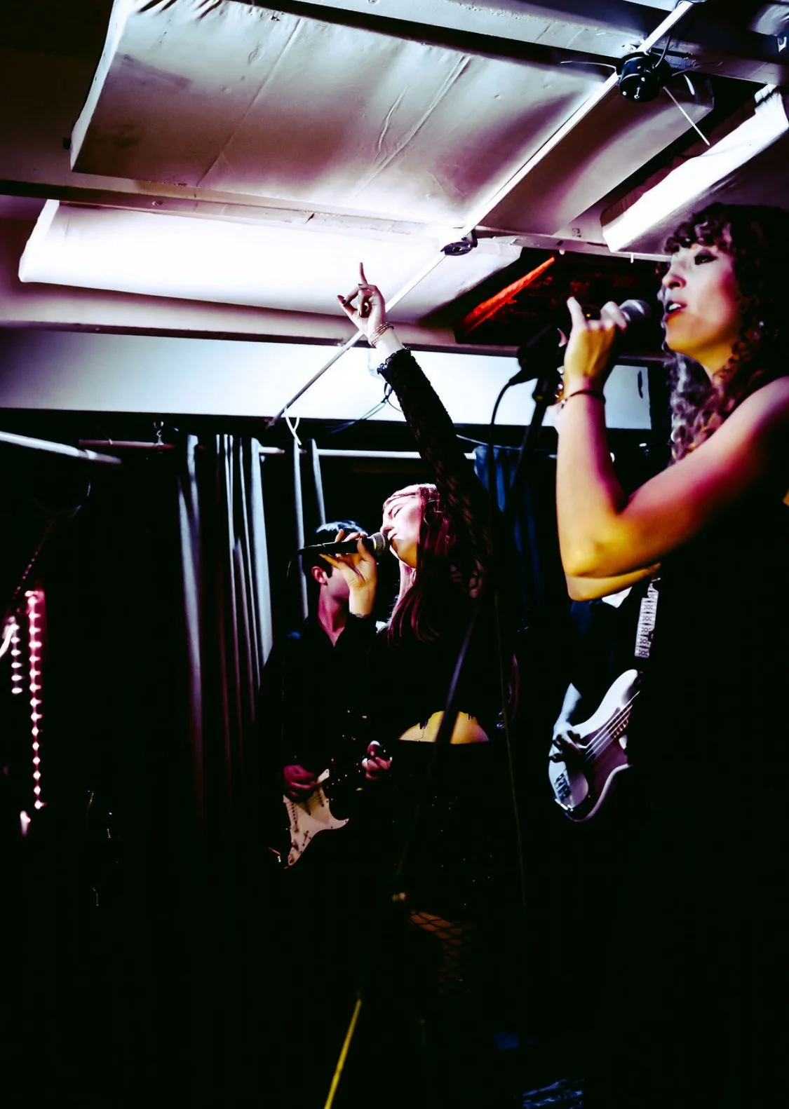Three women performing on stage with microphones and guitars in a dimly lit venue, one woman singing and gesturing with her hand.