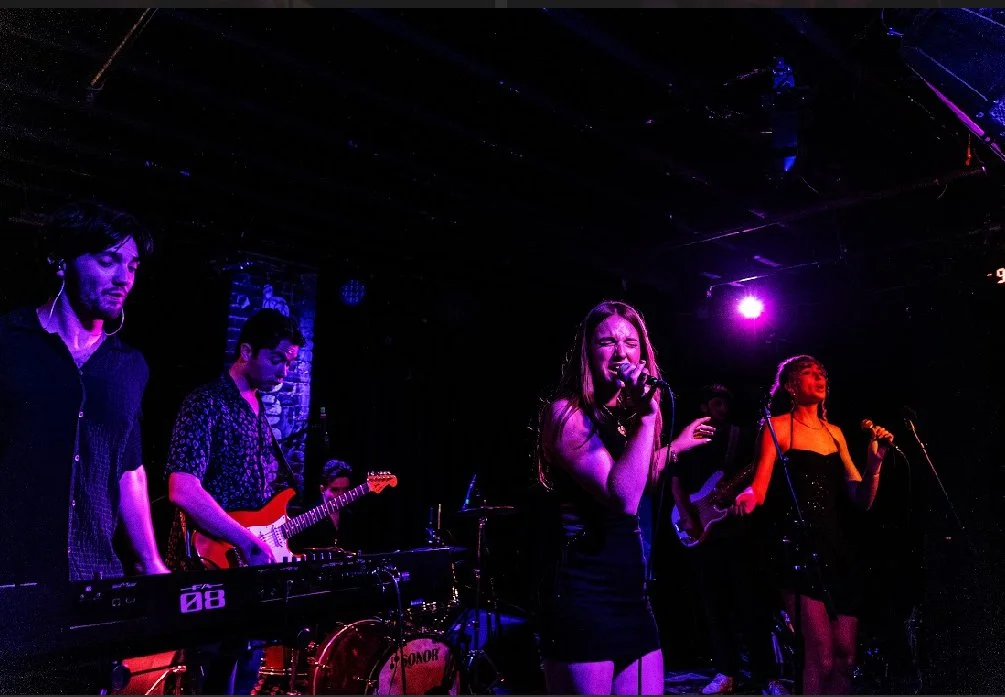 A band performing live on stage with a keyboardist, guitarist, and two female singers under purple stage lighting.