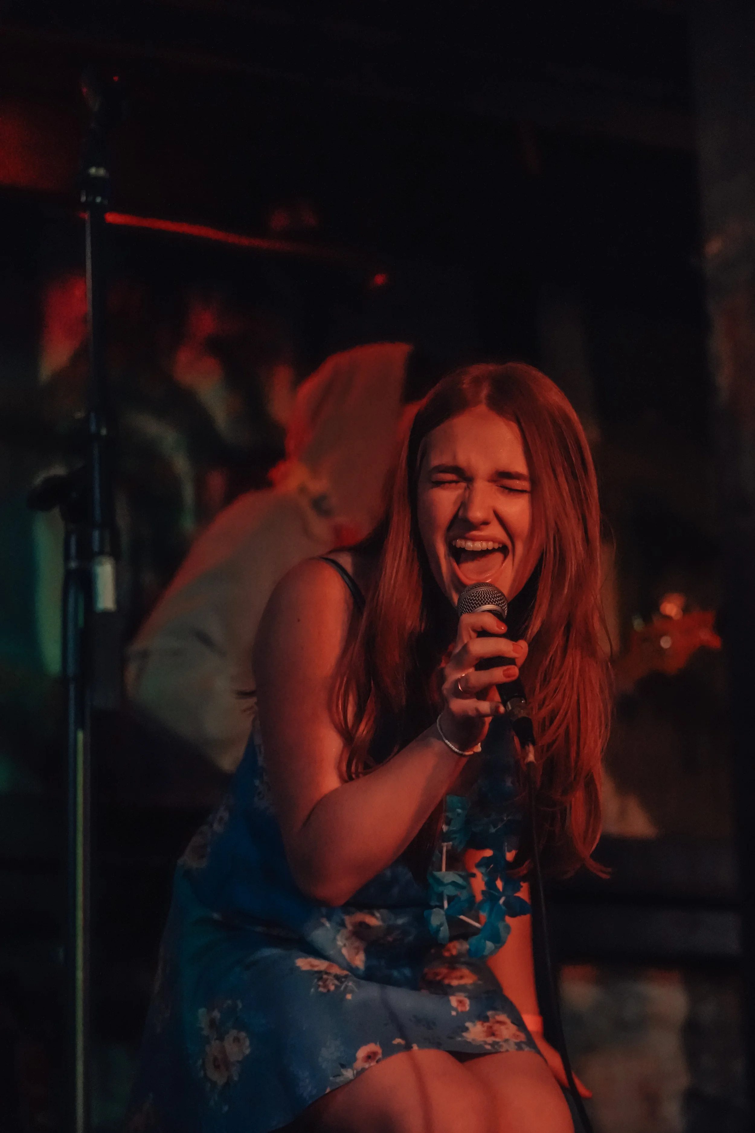 A young woman singing passionately into a microphone on stage, with a woman wearing angel wings in the background.