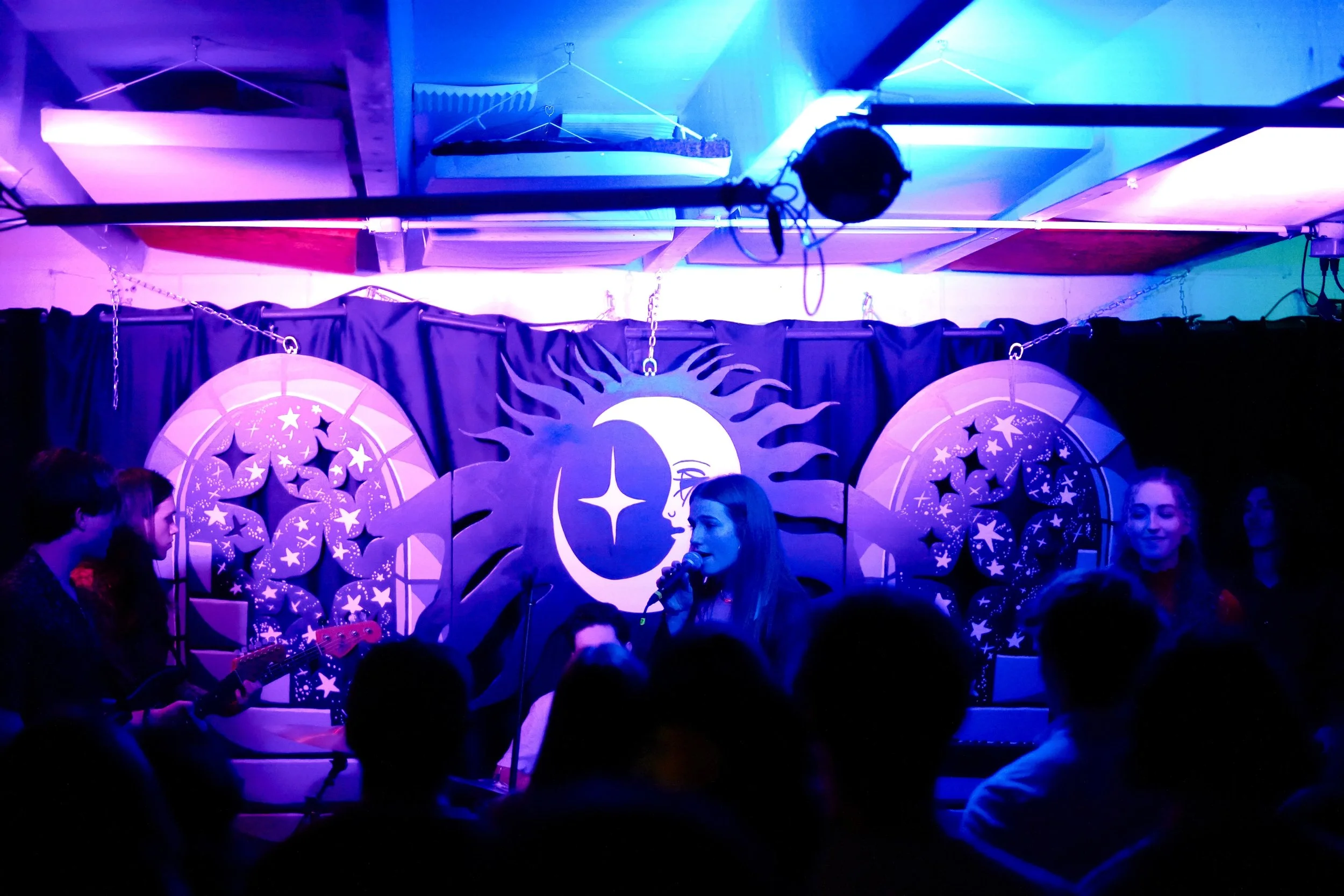 A woman singing into a microphone on stage with a background of moon and star decorations, accompanied by guitar players, under purple and blue stage lighting, with an audience watching