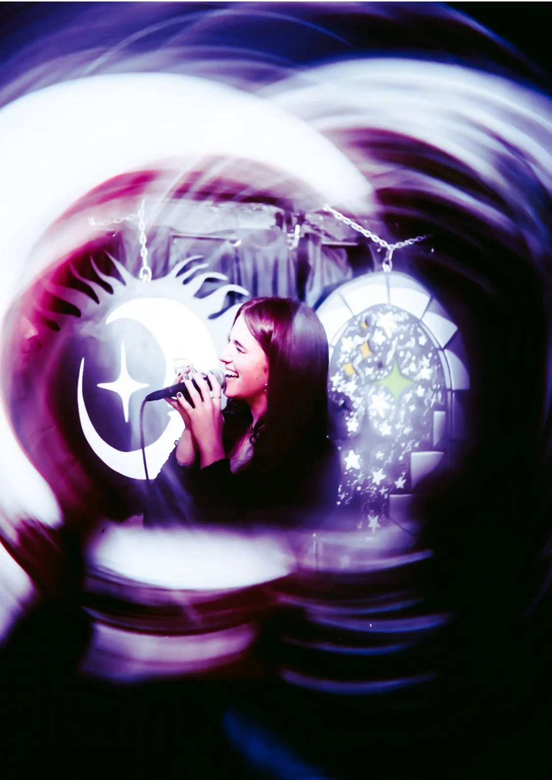 A woman singing into a microphone, with colorful lights and star-themed decorations in the background.