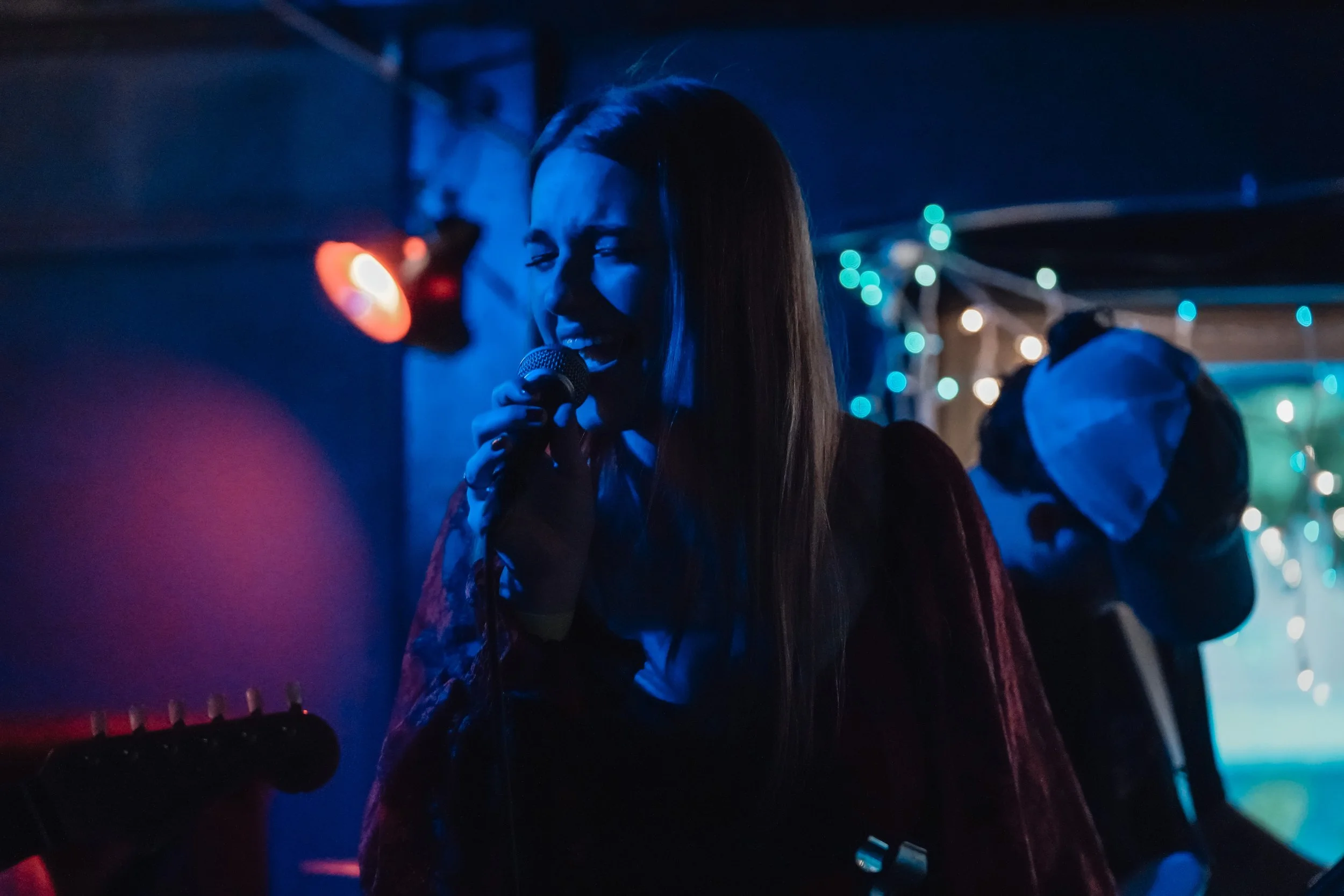 A woman singing into a microphone on stage with colorful lights and a musician in the background.