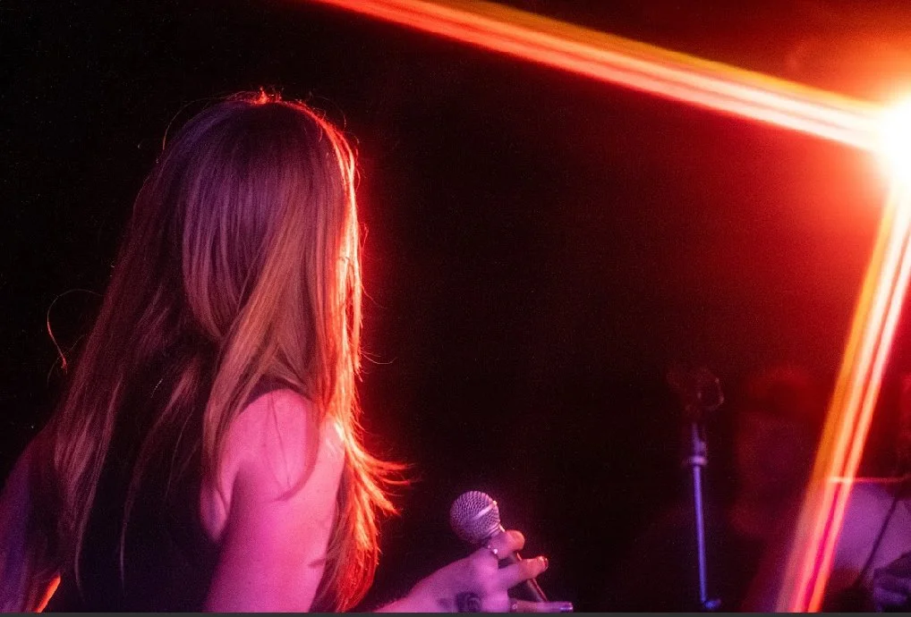 A woman with long hair holding a microphone on stage illuminated by colorful stage lights.