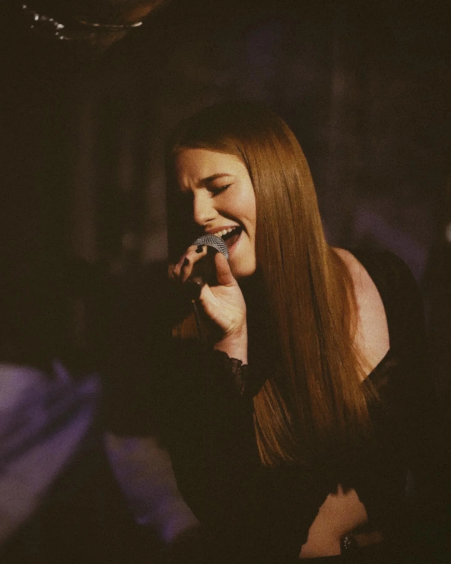 A woman with long red hair singing into a microphone in a dark environment.