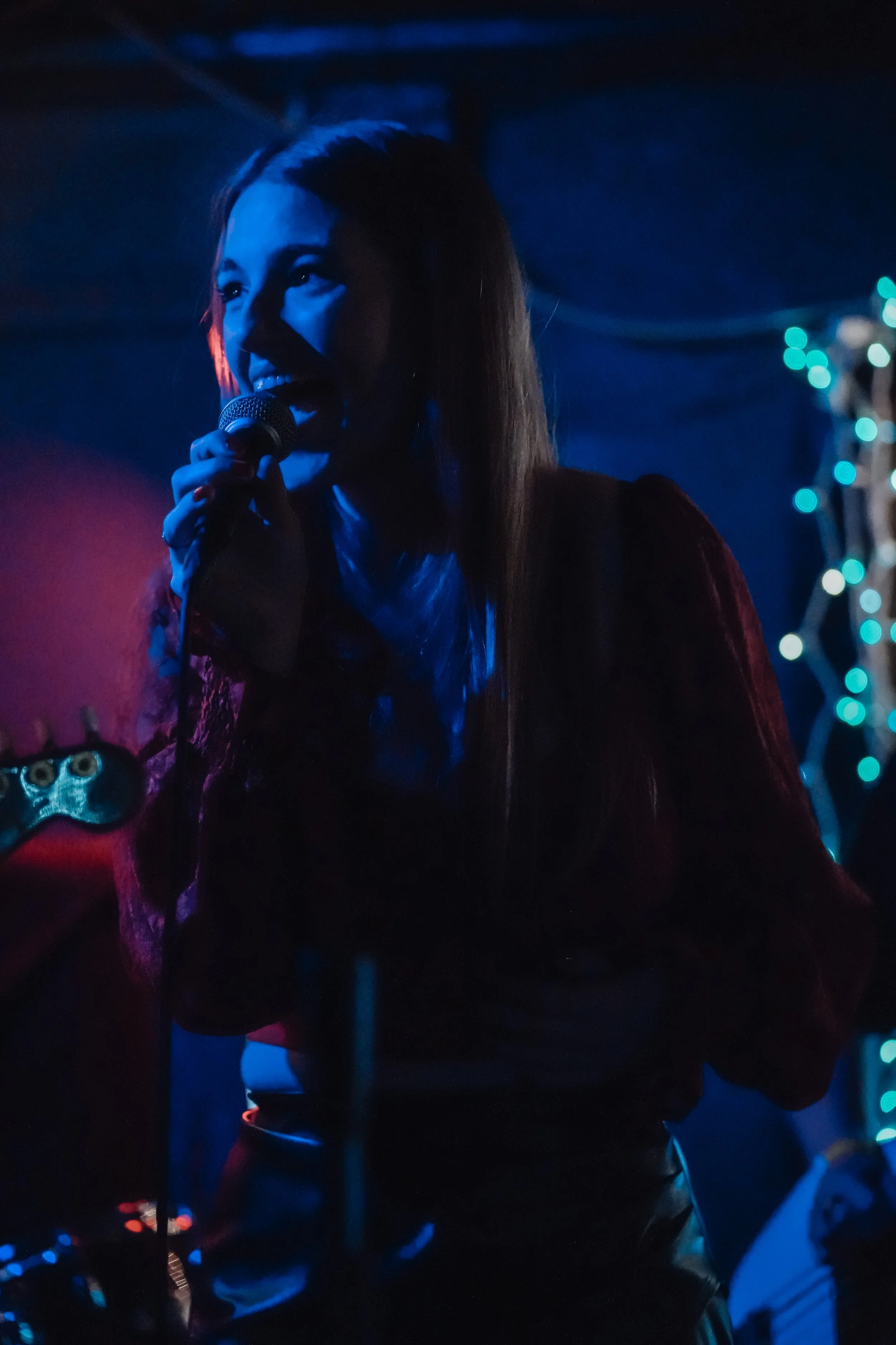 A woman singing into a microphone on stage in a dimly lit environment with blue and red lighting, with blurred decorative lights in the background.