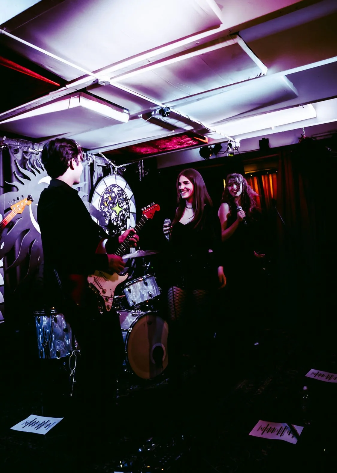 Two young women and three young men performing on a small stage in a dimly lit music venue or club, with one woman holding a microphone, another woman singing, and the man playing an electric guitar, all under colorful stage lighting.