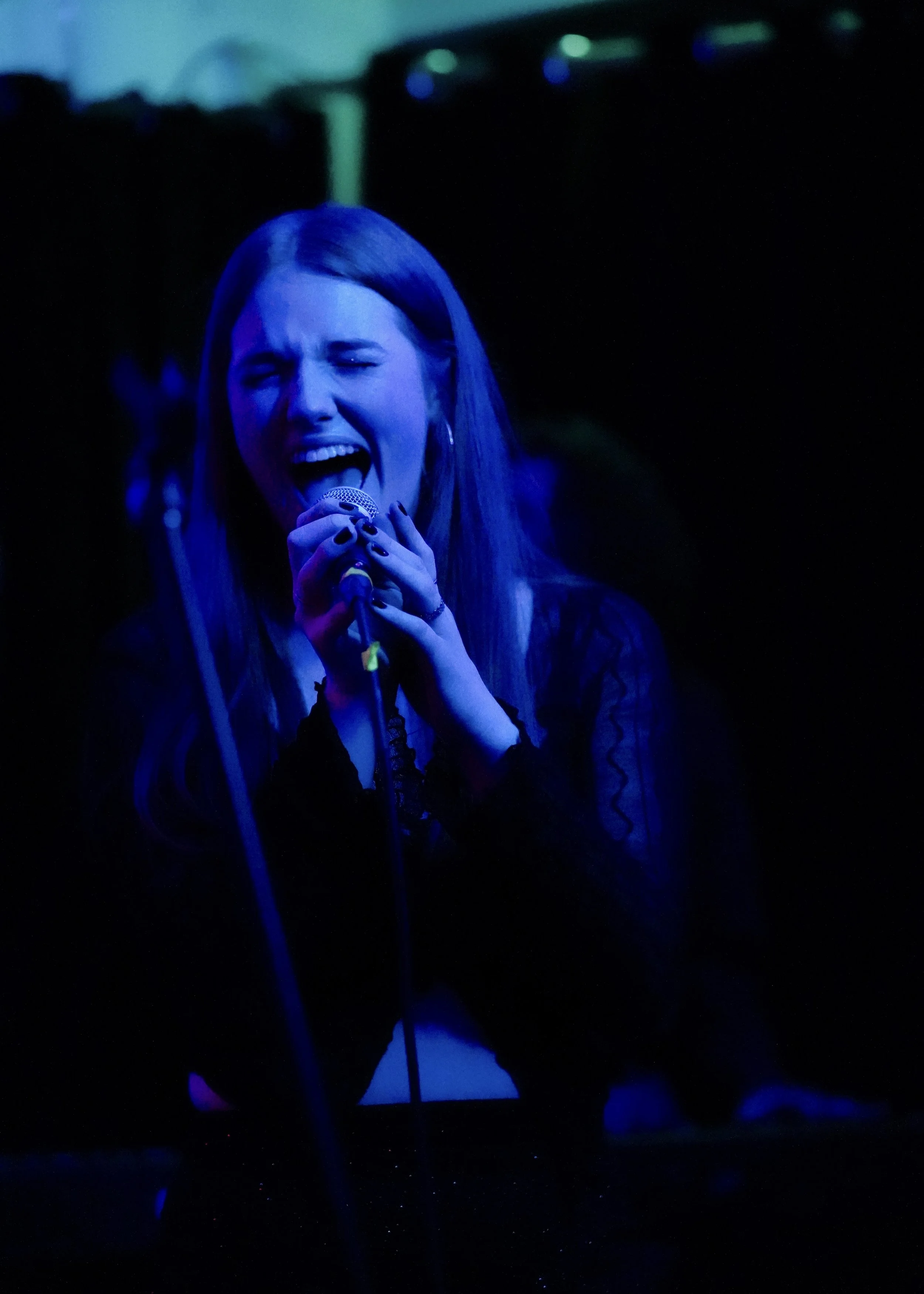 A woman singing passionately into a microphone on stage with dark background and blue lighting.