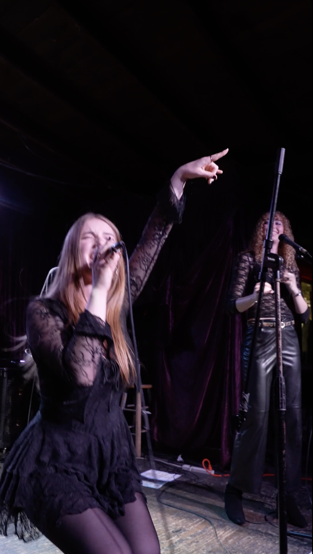 Two women performing on a stage, one singing into a microphone with her eye closed and hand pointing upwards, both wearing black lace outfits.