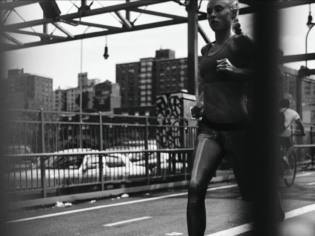 A woman jogging outdoors on a city bridge or overpass with high-rise buildings in the background in black and white.