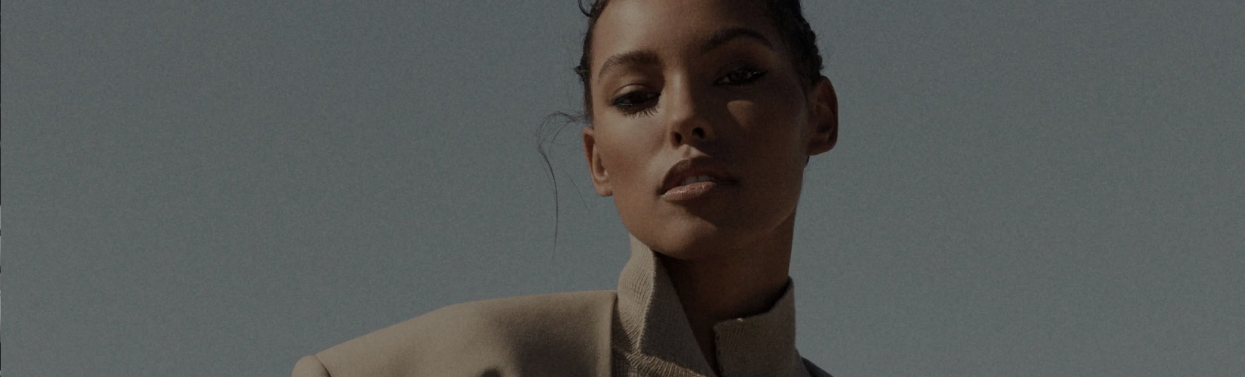 Close-up of a woman with dark hair styled in braids, wearing a beige blazer, against a clear sky background.