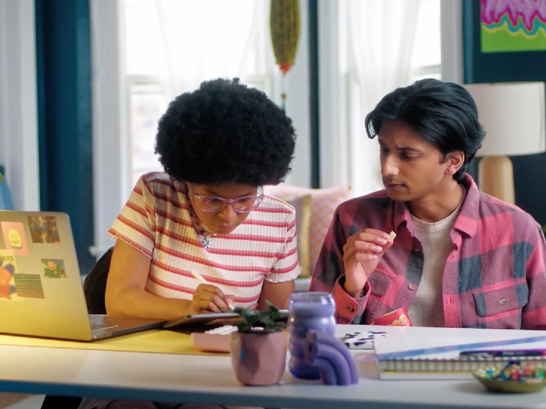 Two people sitting at a table, one of them looking at a notebook or tablet, the other holding a snack, with a laptop, colorful decorations, and a potted plant on the table.