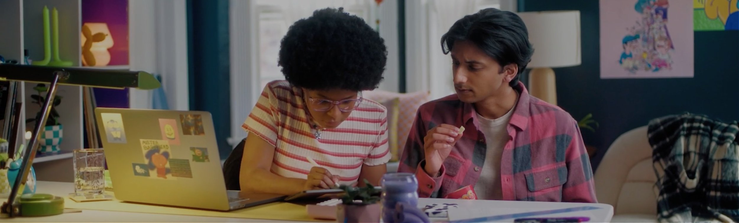 A young woman and a young man are sitting at a table, looking at a tablet device together. The woman has glasses and curly hair, and the man has dark wavy hair. The table has various items including a laptop, a jar, and some papers. The background shows a colorful room with bookshelves, artwork, and a window.