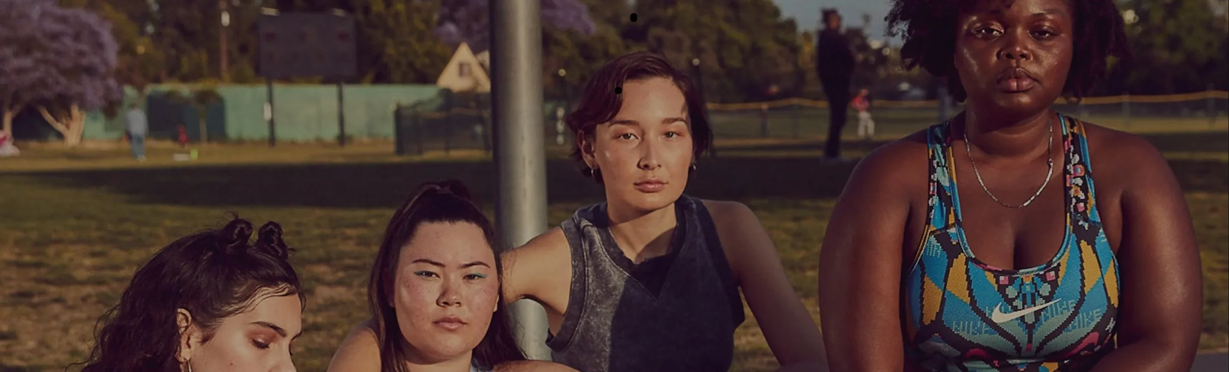 Four women sitting outdoors in a park on a sunny day, with trees, a fence, and people in the background.