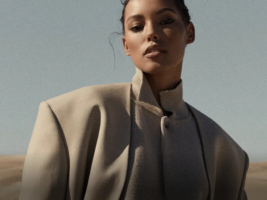 A woman with dark hair pulled back, wearing a beige jacket and a beige high-neck top, looking at the camera with a neutral expression against a clear sky background.