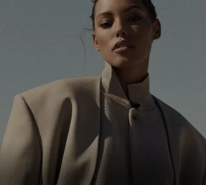 A woman wearing a beige jacket with a high collar, looking slightly downward against a plain background.