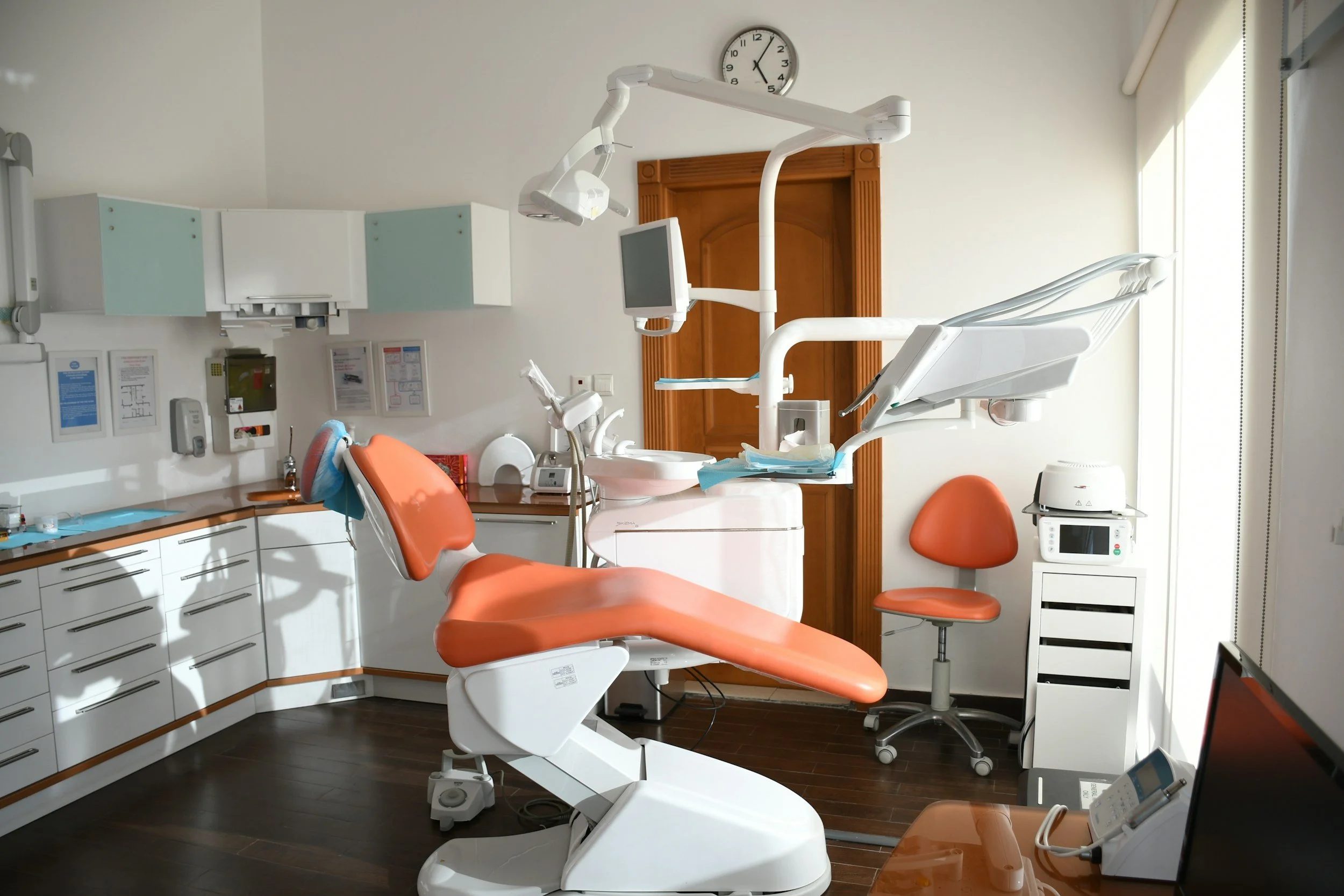 Empty dental examination room with an orange dental chair, dental equipment, and a computer in the corner.