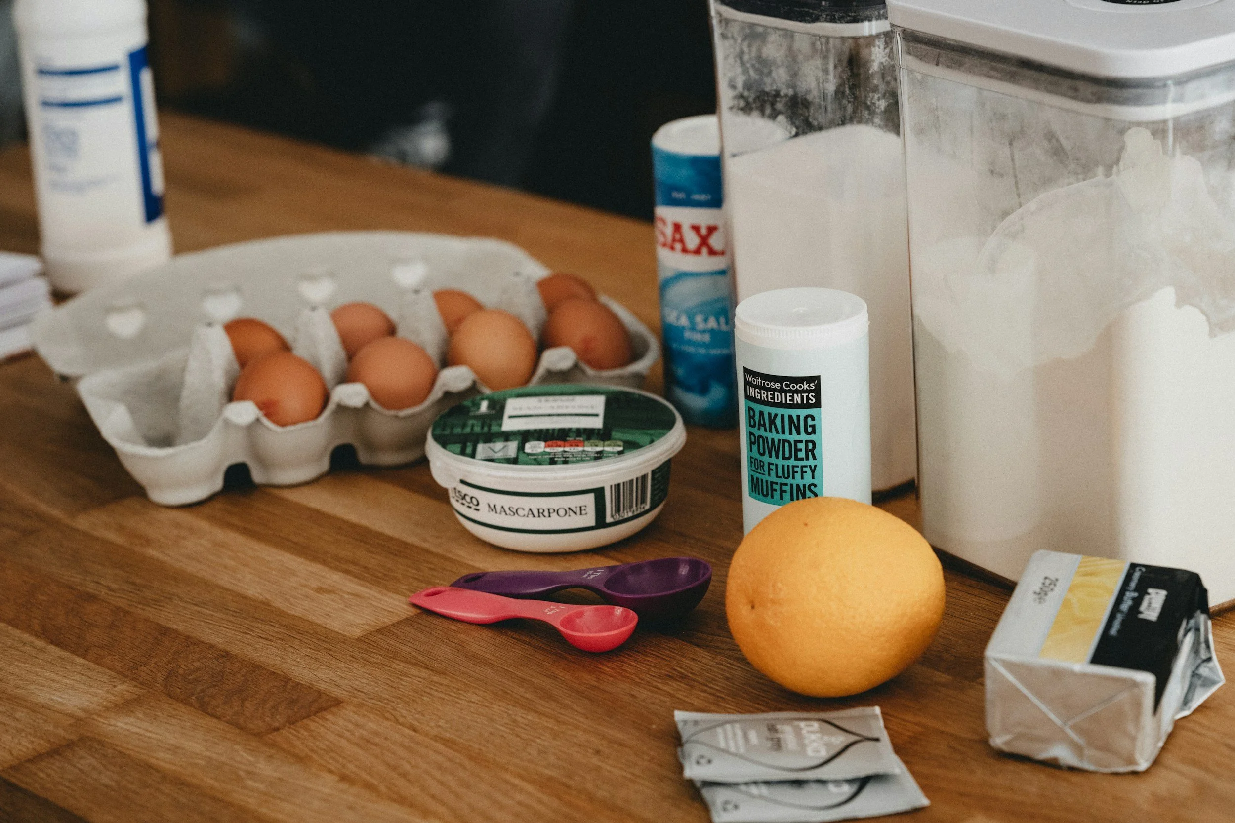 A wooden countertop with various baking ingredients and utensils, including a carton of eggs, a tub of mascarpone cheese, a container of baking powder, a lemon, a packet of butter, a small pink and purple measuring spoon, and a white storage container.