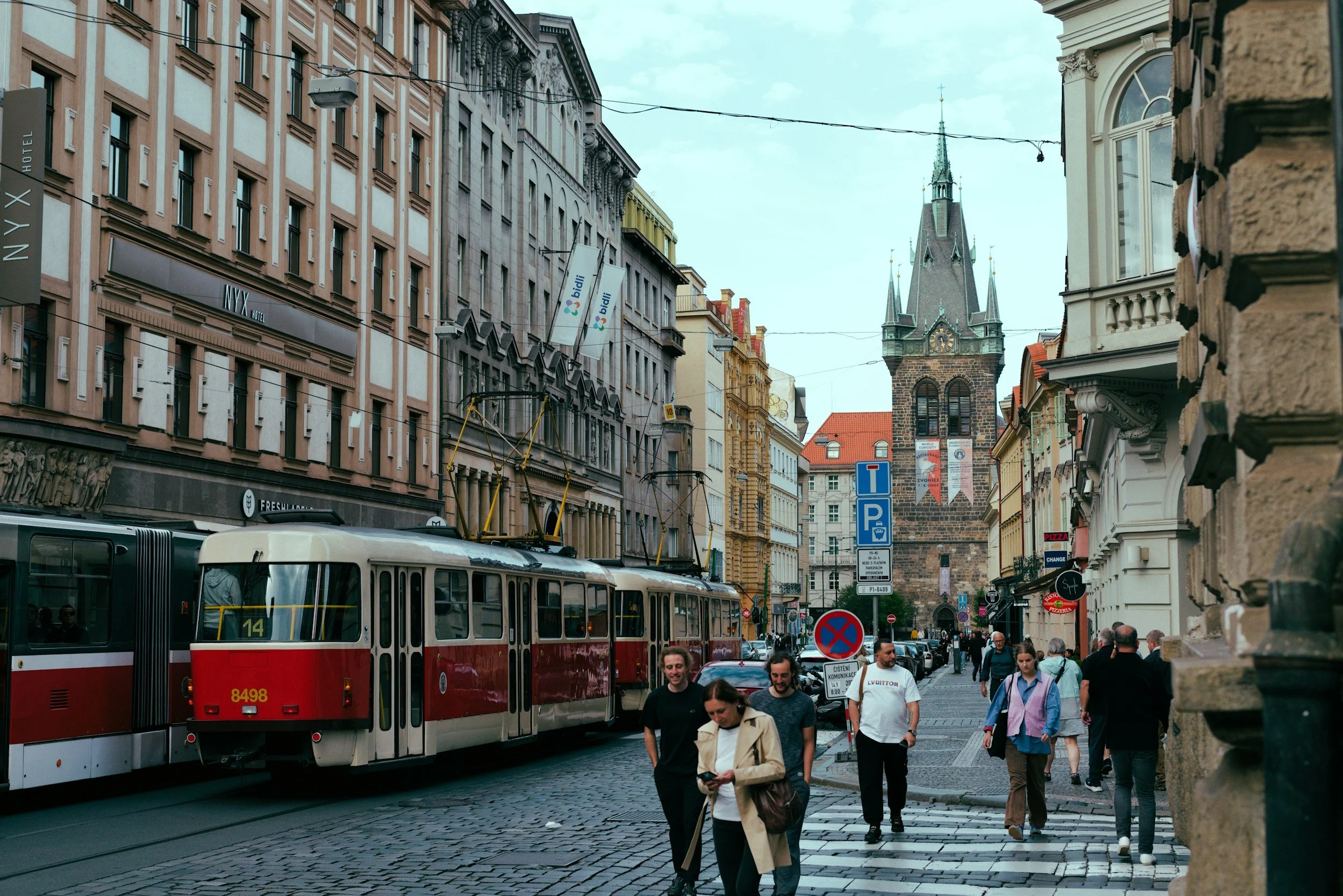 A busy street scene in a European city, with a tram, pedestrians walking, historic buildings, and a church steeple in the background.