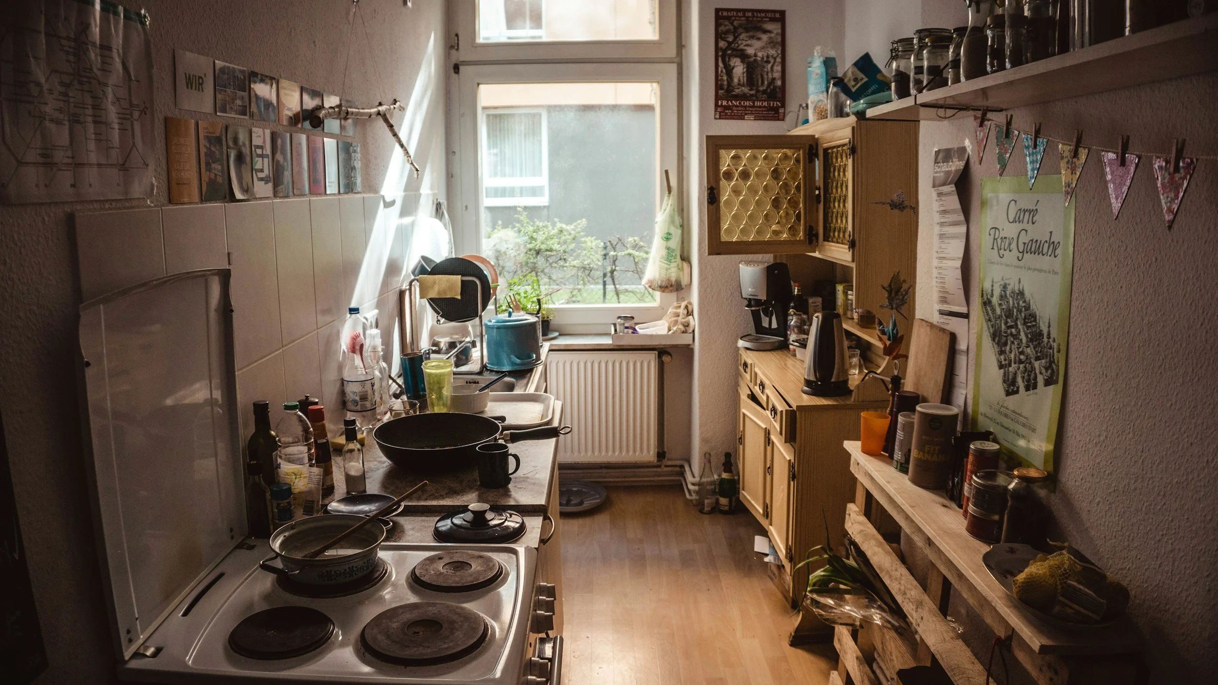 Cluttered kitchen with dishes, bottles, and cookware on the counter, a window with sunlight, and shelves holding jars and items, with a wooden pallet shelf on the right, and wall decorations including a poster and colorful bunting.