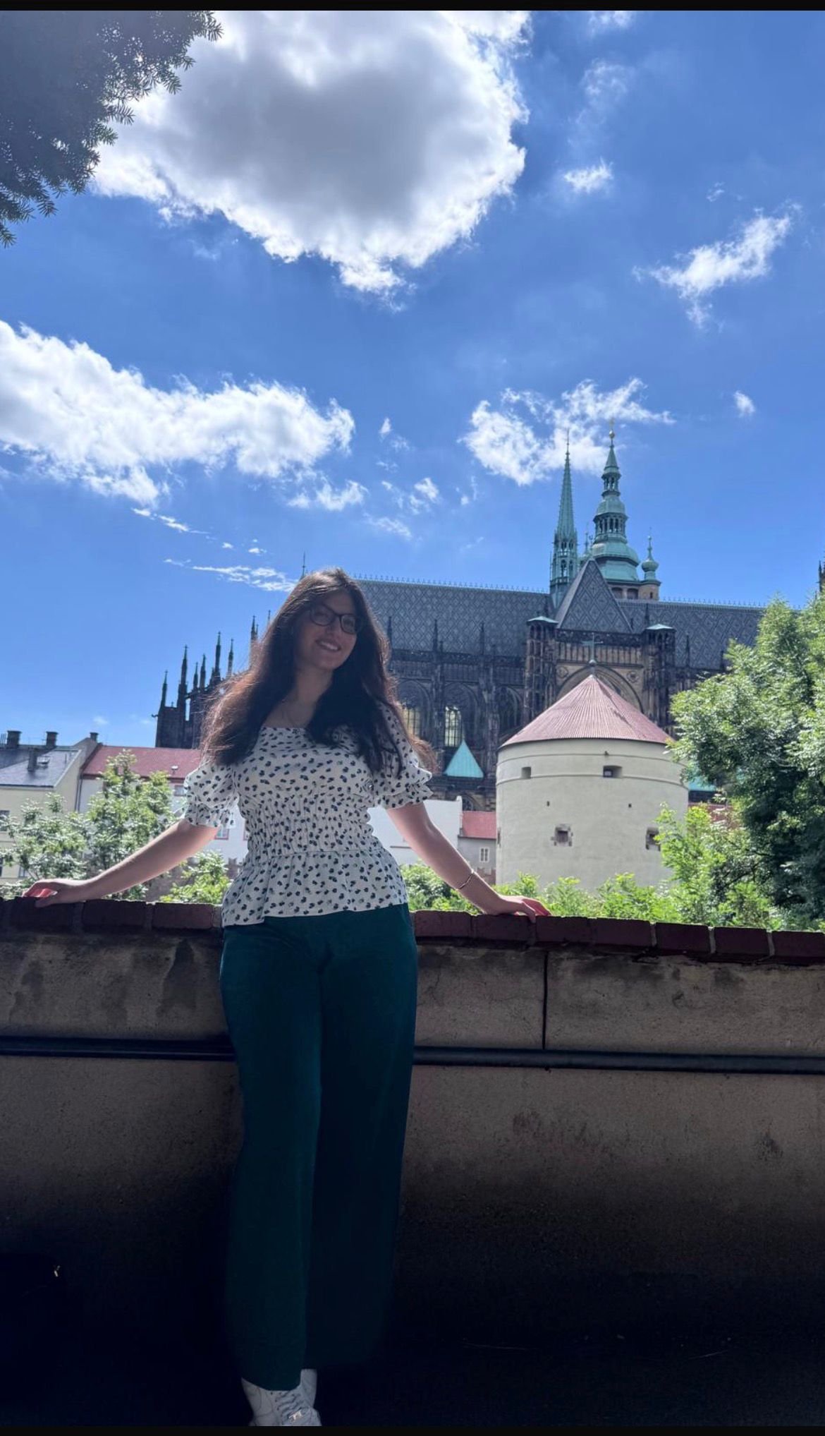 A woman with long hair and glasses standing outdoors, leaning against a brick-lined wall, with a historic cathedral in the background under a partly cloudy blue sky.