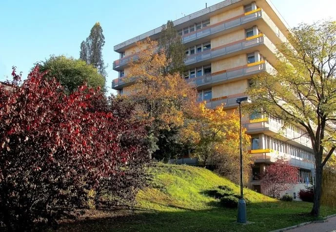Multi-story residential building with balconies, surrounded by colorful autumn trees and greenery under a clear blue sky.