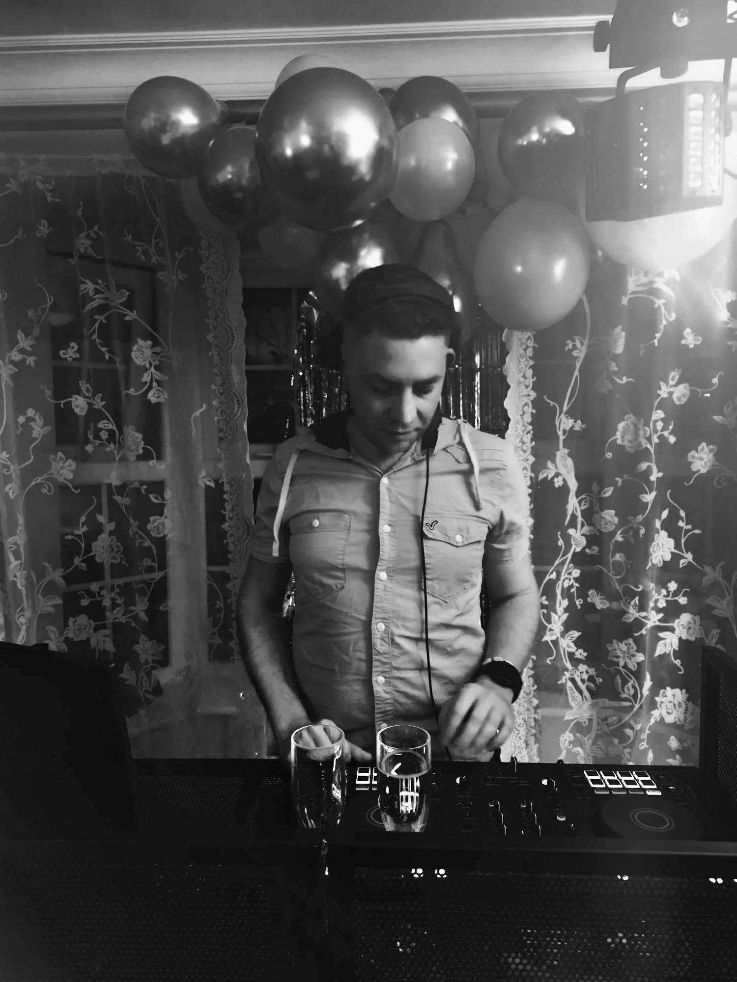 A man with headphones standing behind a DJ console, with two glasses of drink in front of him, at a celebration party with balloons and decorative curtains in the background.