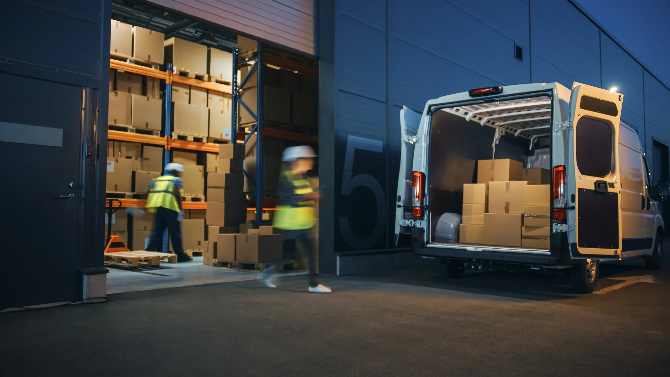 Workers loading and unloading boxes from a delivery van outside a warehouse at night.