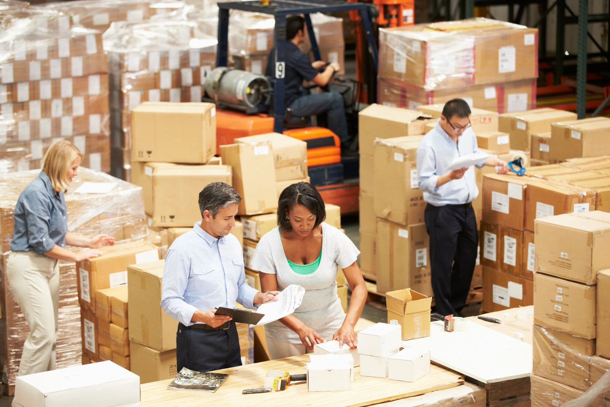 Workers in a warehouse managing and inspecting packages surrounded by stacked boxes and pallets, with a forklift in the background.