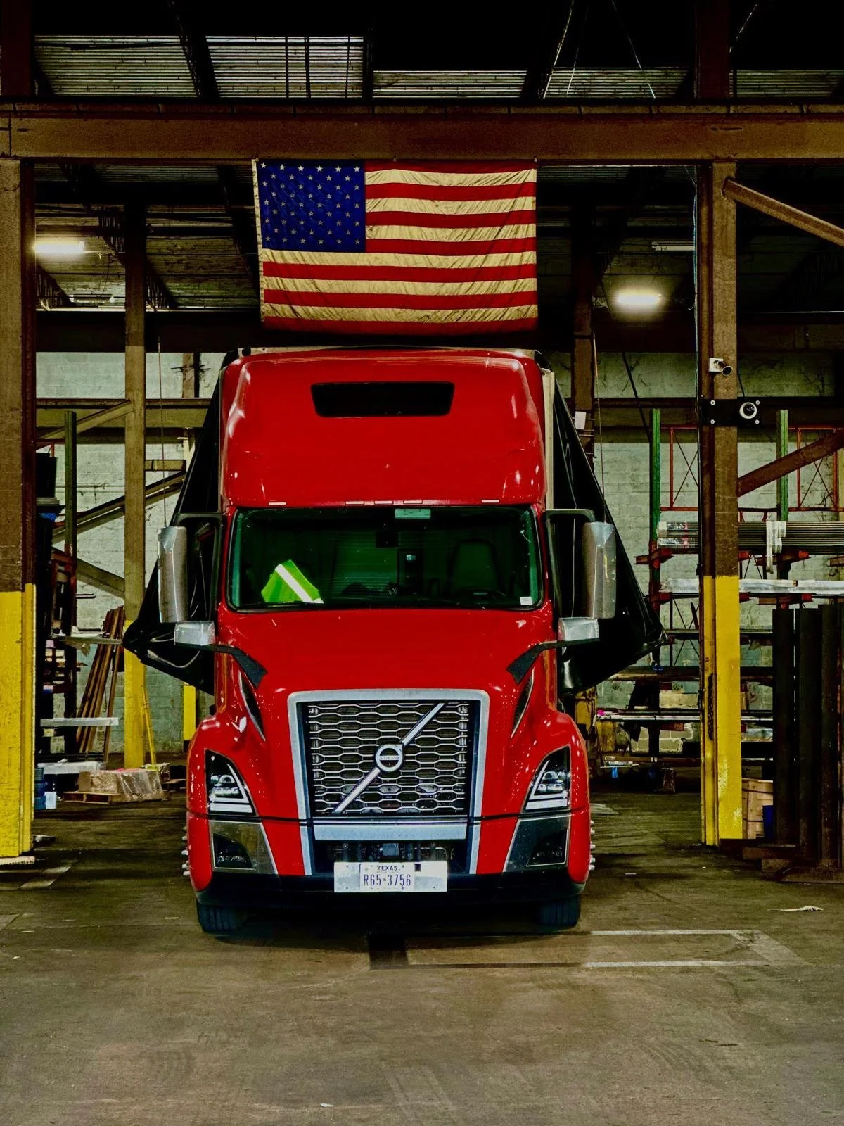 Red semi-truck parked inside a warehouse or garage with an American flag hanging overhead.