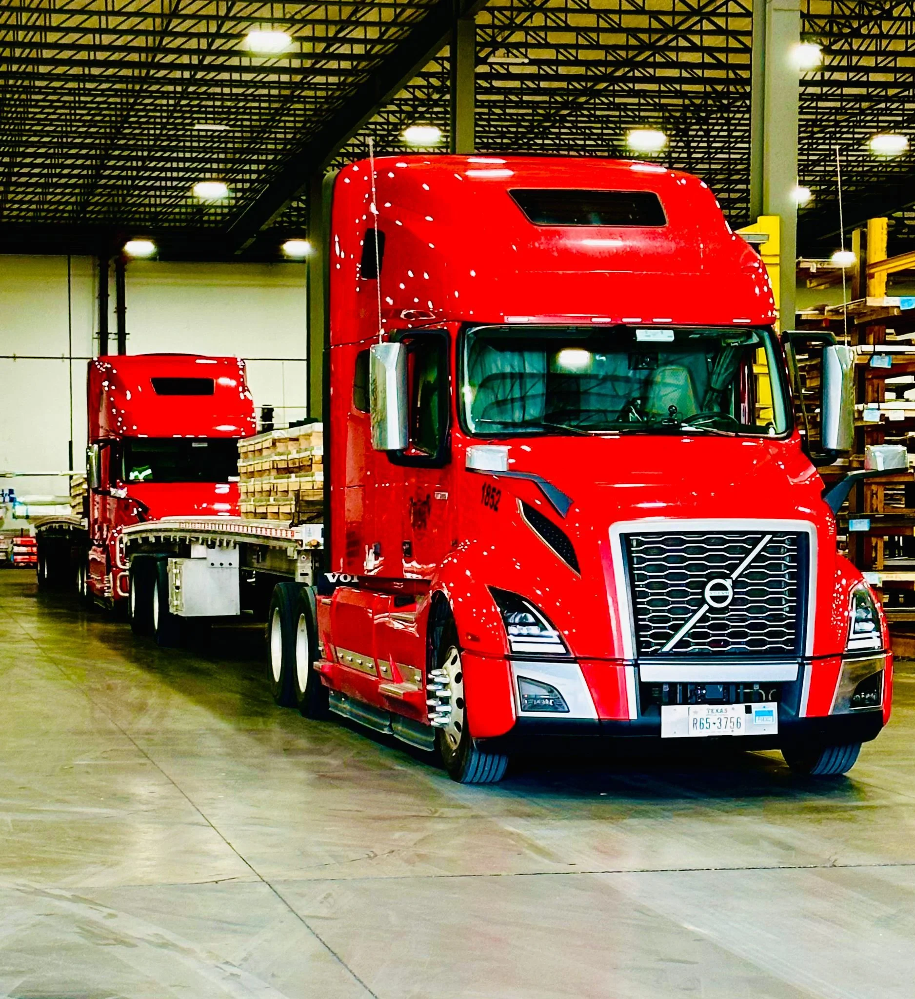 Red semi-truck inside a warehouse with shelving on the right and stacked wood on the trailer.