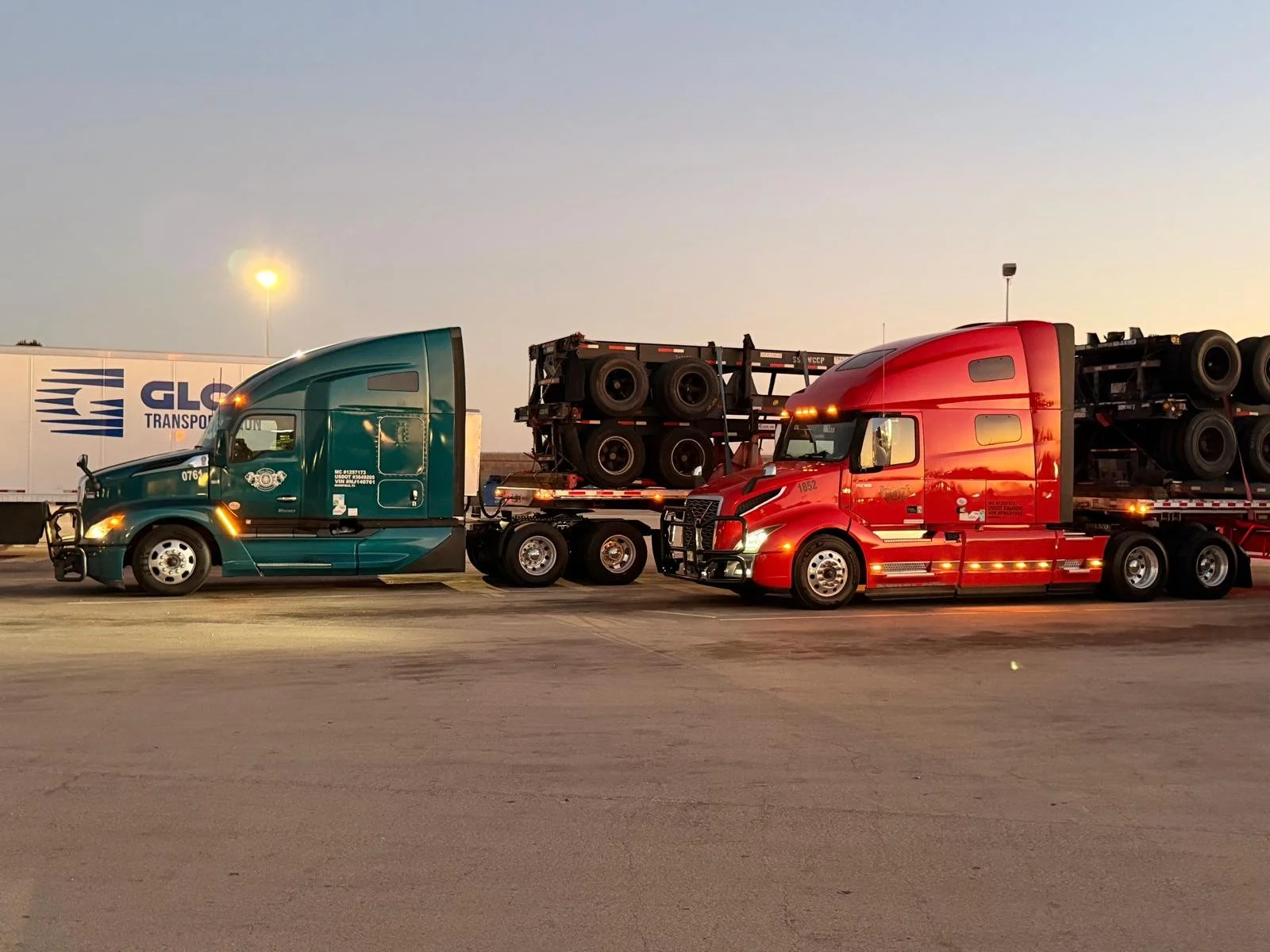Two semi trucks, one green and one red, parked side by side at dusk. Both trucks are hauling multiple tires on their flatbeds.