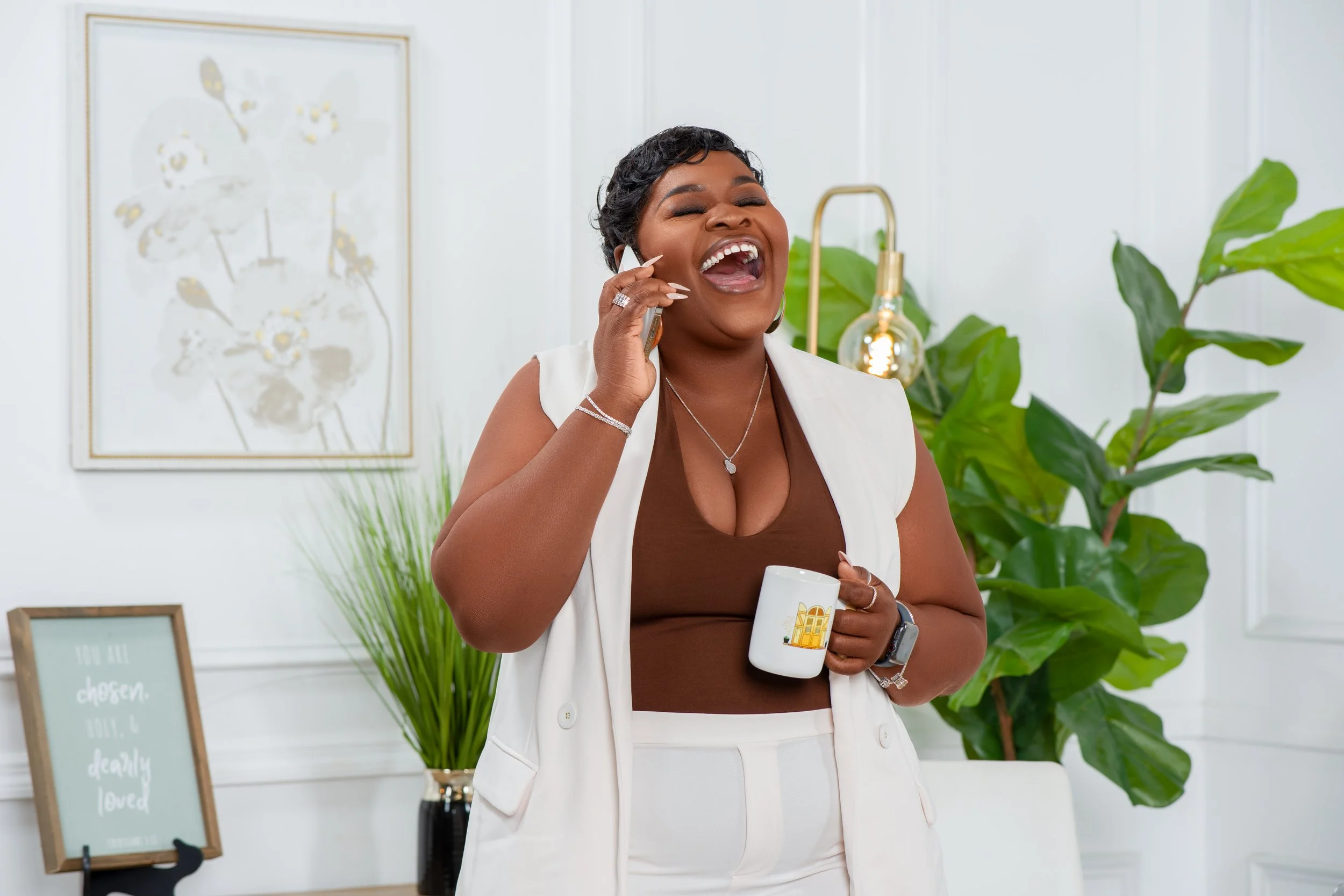 A woman with short black hair, wearing a white vest, brown top, and white pants, smiling and talking on her phone while holding a white mug. She is standing in a bright, modern room with green plants and framed wall decor.