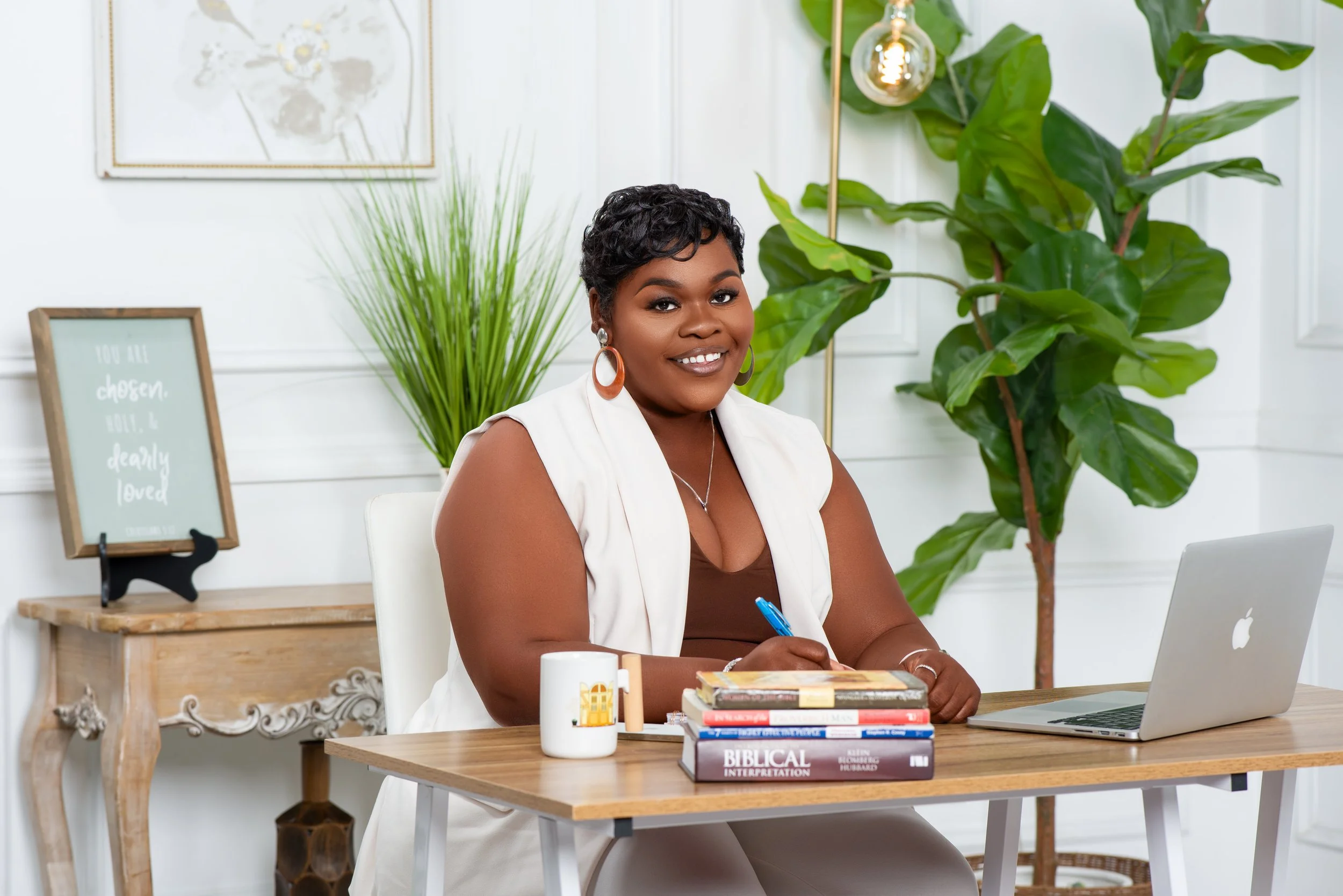 A woman sitting at a desk with a smile, wearing a white vest and earrings, with a laptop, a mug, and a stack of books including one titled 'Biblical Interpretation' in a bright, modern office space with green plants and framed wall art.