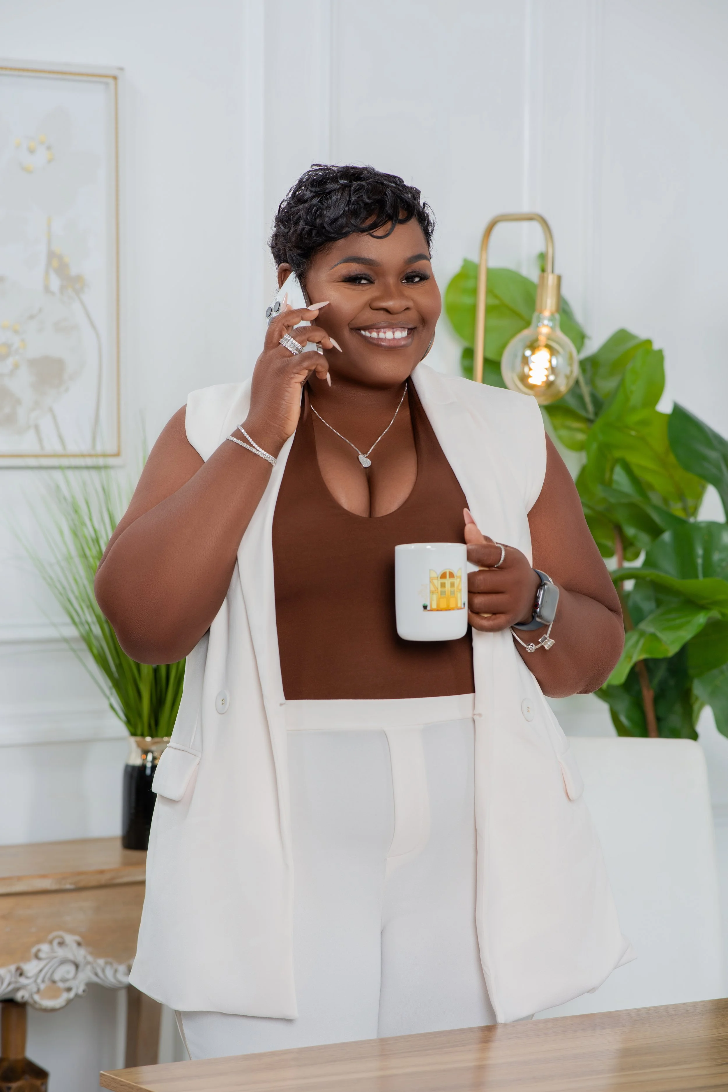 A smiling woman with short dark curly hair talking on a cell phone while holding a white mug with a yellow building illustration. She is wearing a brown top and a light-colored vest and pants, and is accessorized with jewelry and a smartwatch. The background includes a large green plant, a modern lamp, and wall art.