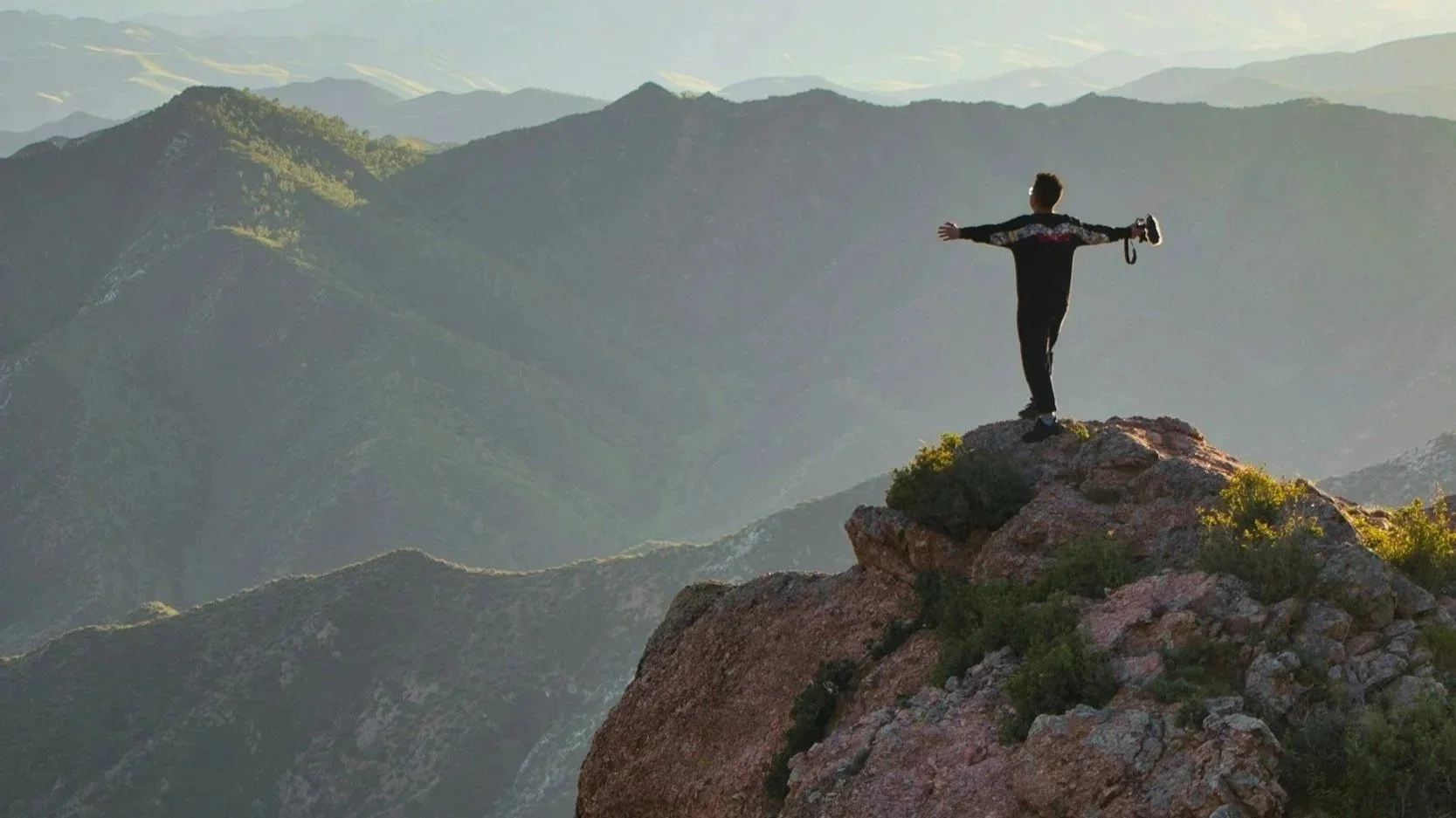 Person standing on a mountain peak with arms outstretched overlooking a range of mountains.
