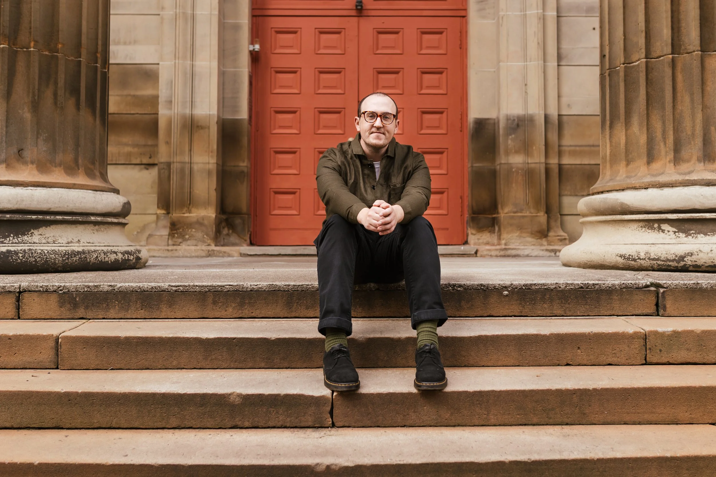 A man with glasses sitting on the steps in front of a red wooden door surrounded by stone columns.