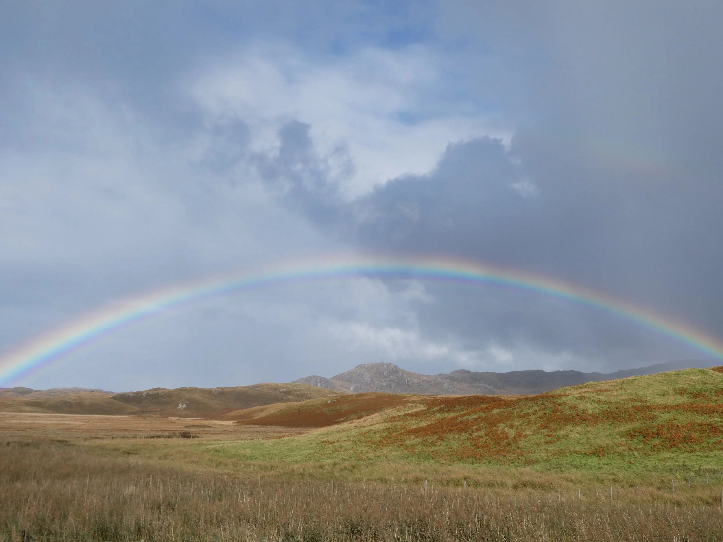 Scenic landscape with rolling grassy hills, mountains in the distance, a partly cloudy sky, and a rainbow arching across the sky.