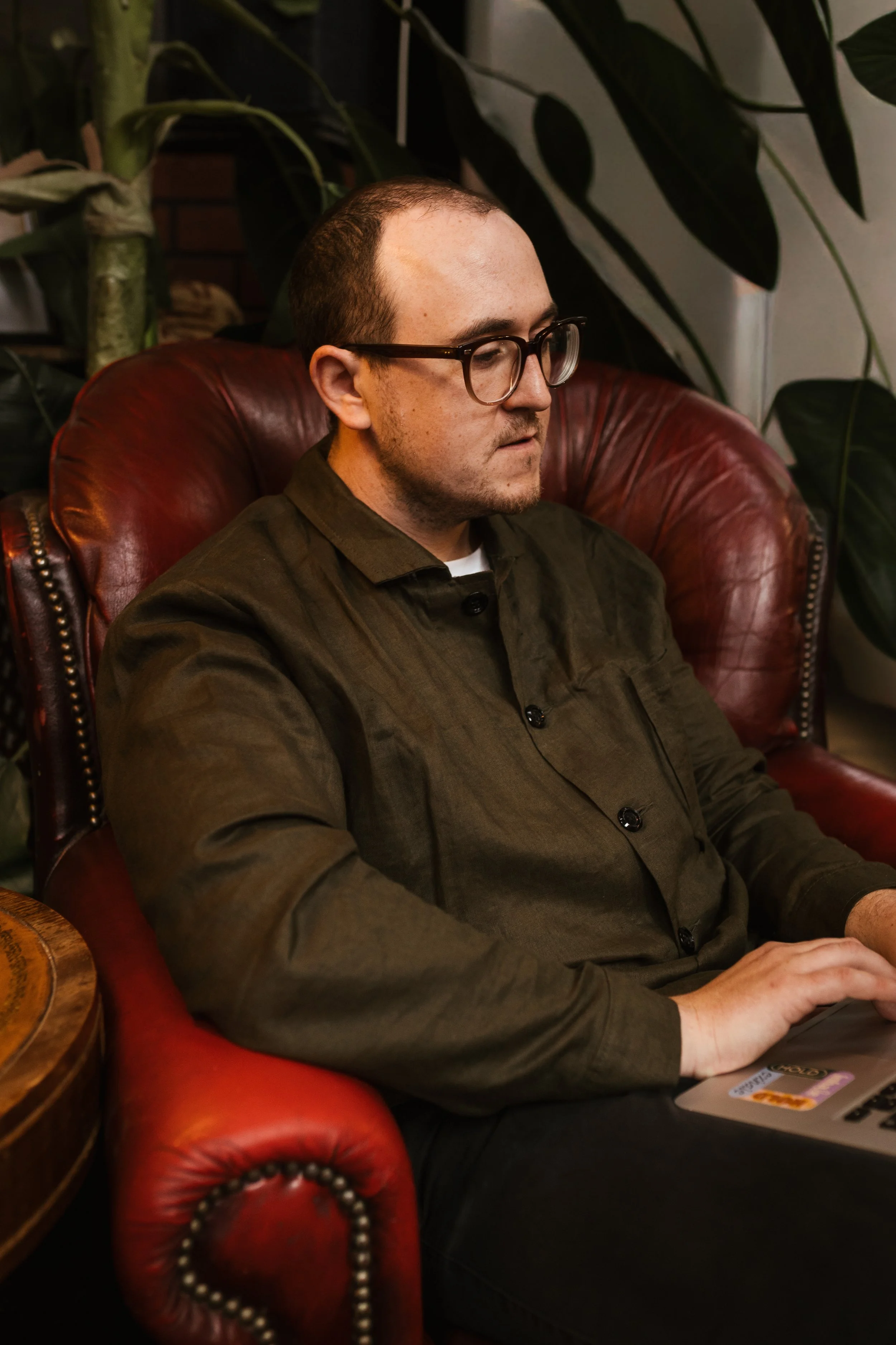 A man with glasses sitting on a red leather armchair, working on a laptop in a cozy indoor setting with large leafy plants behind him.
